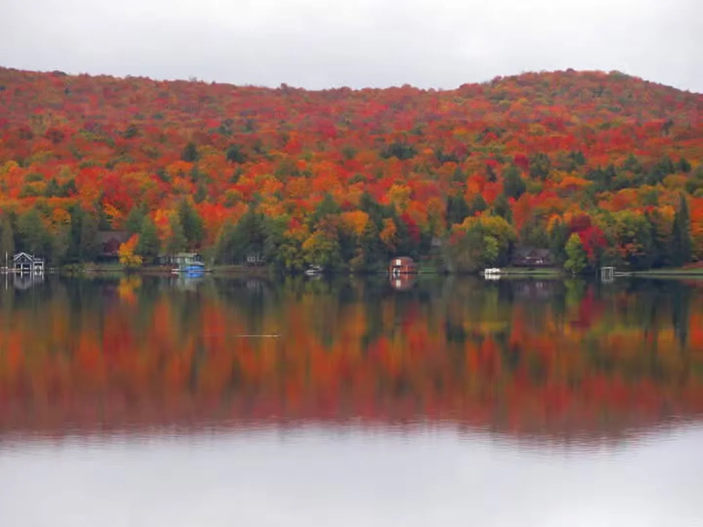 A lake reflecting a hillside covered with colorful autumn foliage including red, orange, and yellow trees, with a cloudy sky overhead.