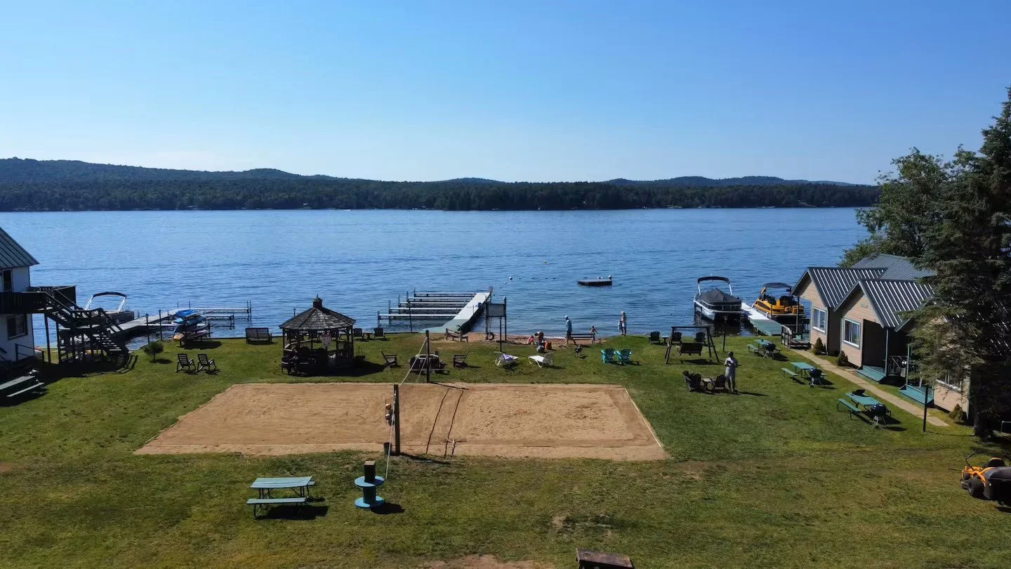 A lakeside scene with houses, a sandy volleyball court, boats docked by the shore, and people enjoying the sunny day.