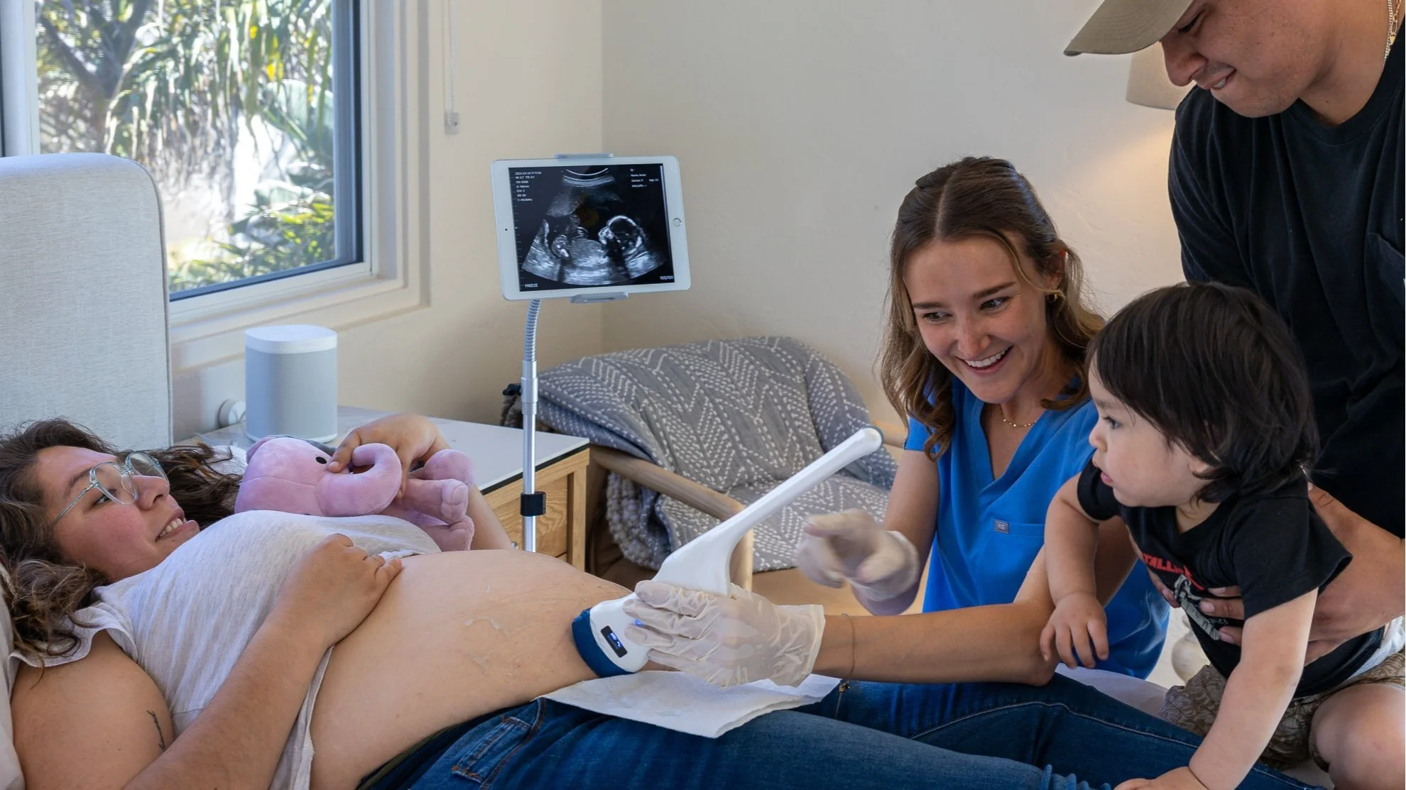 A woman lying in a hospital bed during an ultrasound with a pink stuffed animal, surrounded by a woman in blue scrubs, an older man, and a young boy in a room with a window, ultrasound scan on a monitor, and medical equipment.