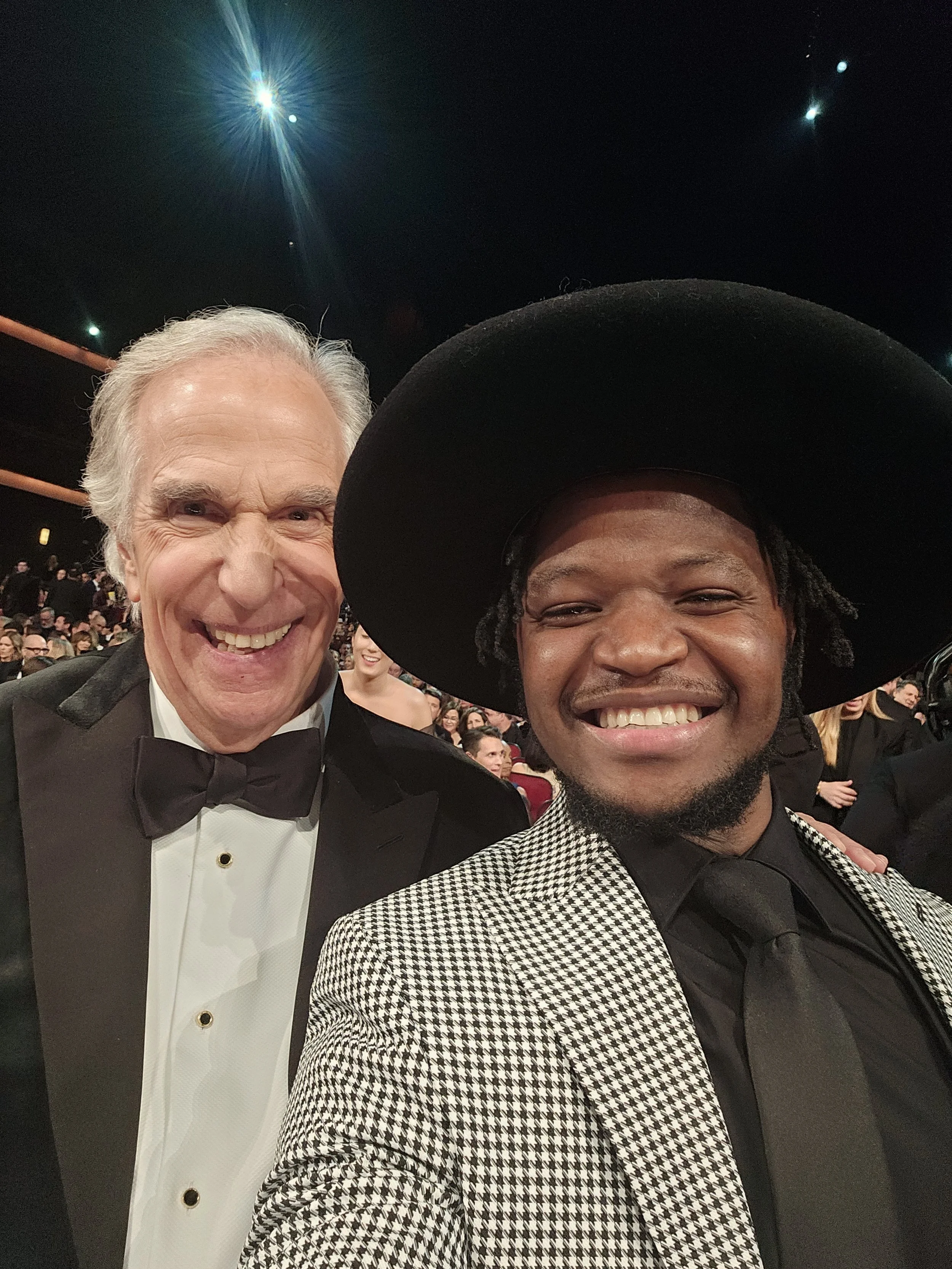 Two men dressed in tuxedos taking a selfie at an event, with a large crowd in the background and bright stage lights overhead.