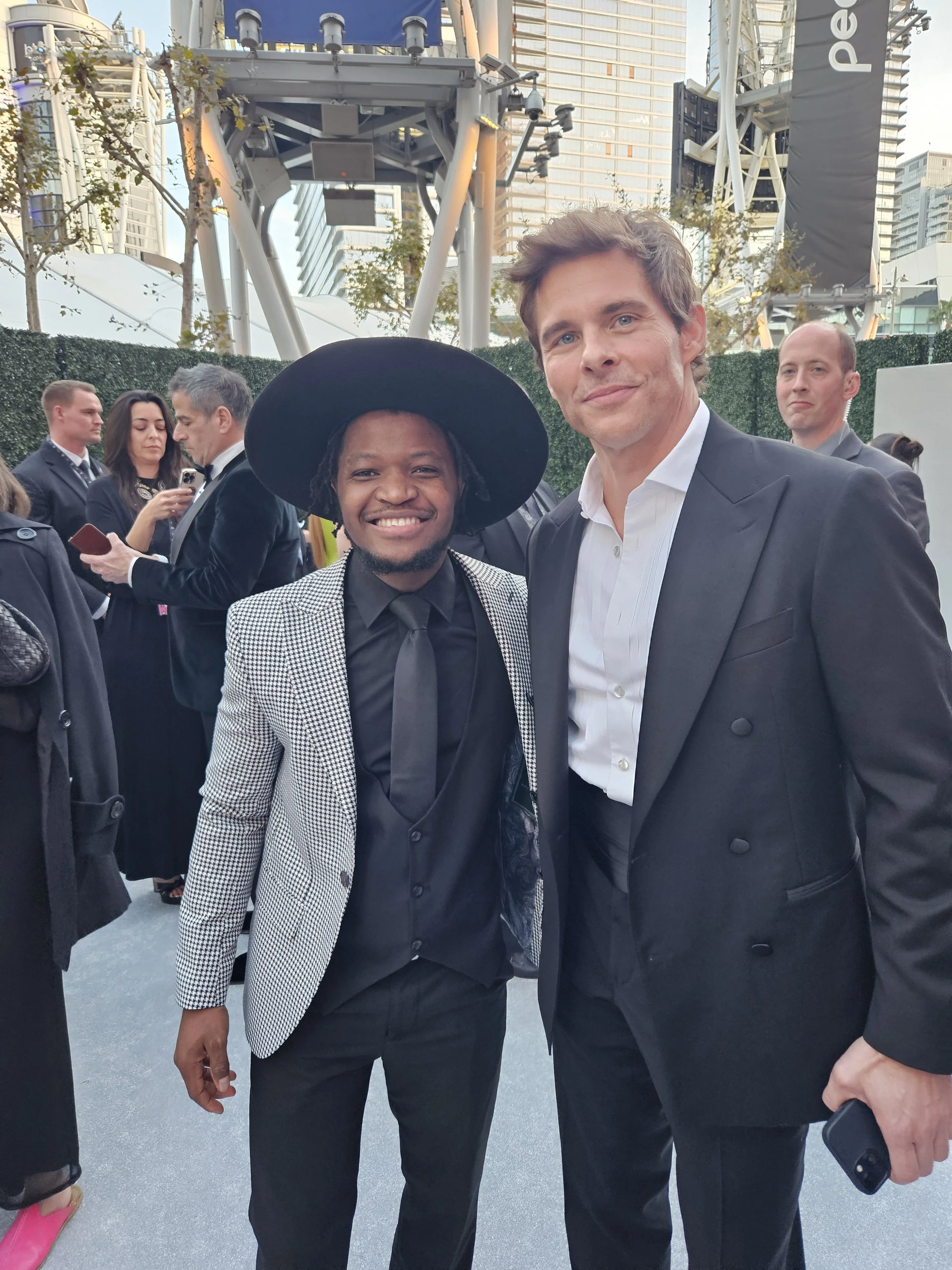 Two men in formal attire smiling at an outdoor event in a city with skyscrapers, cameras, and people in the background.