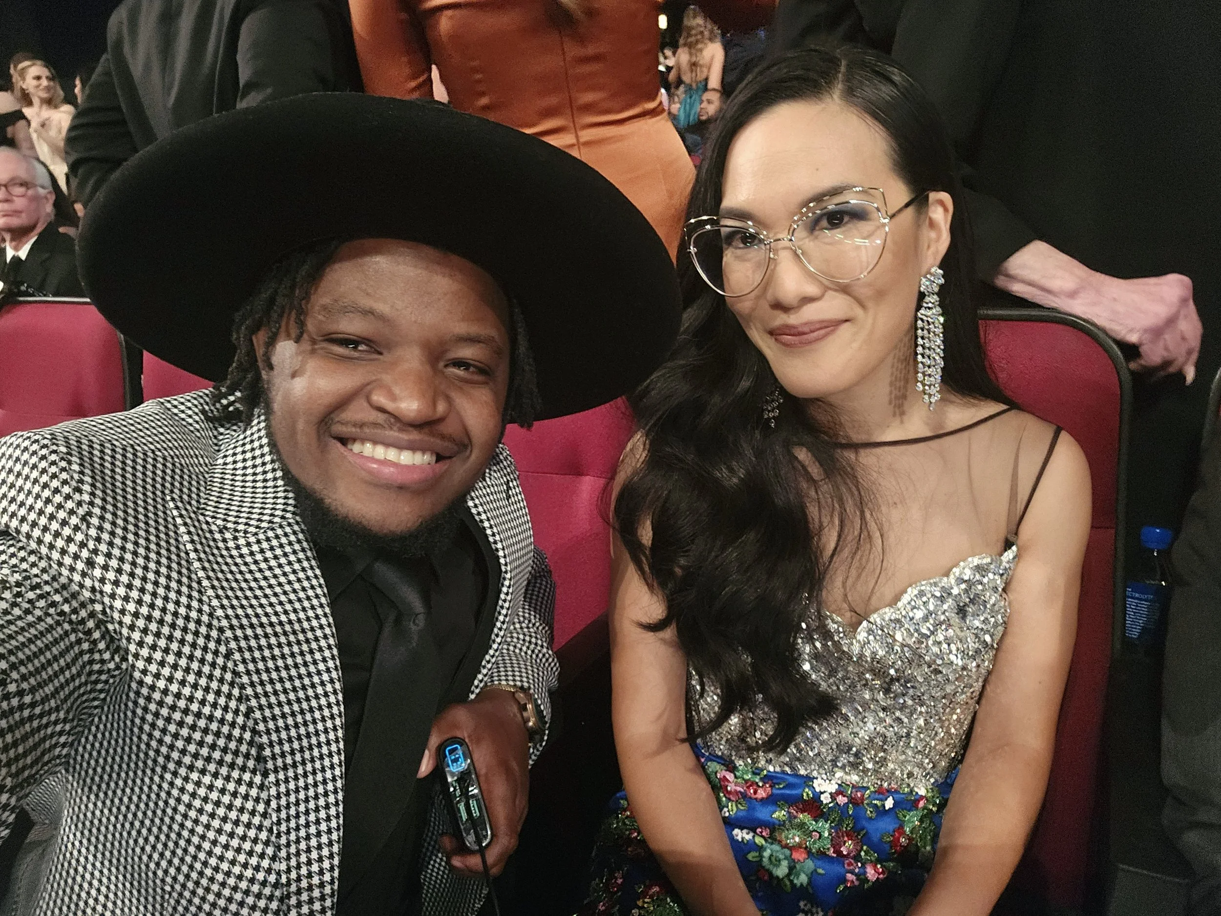 Two people sitting at an event, smiling at the camera, with a woman wearing glasses and a woman in a sparkly dress.