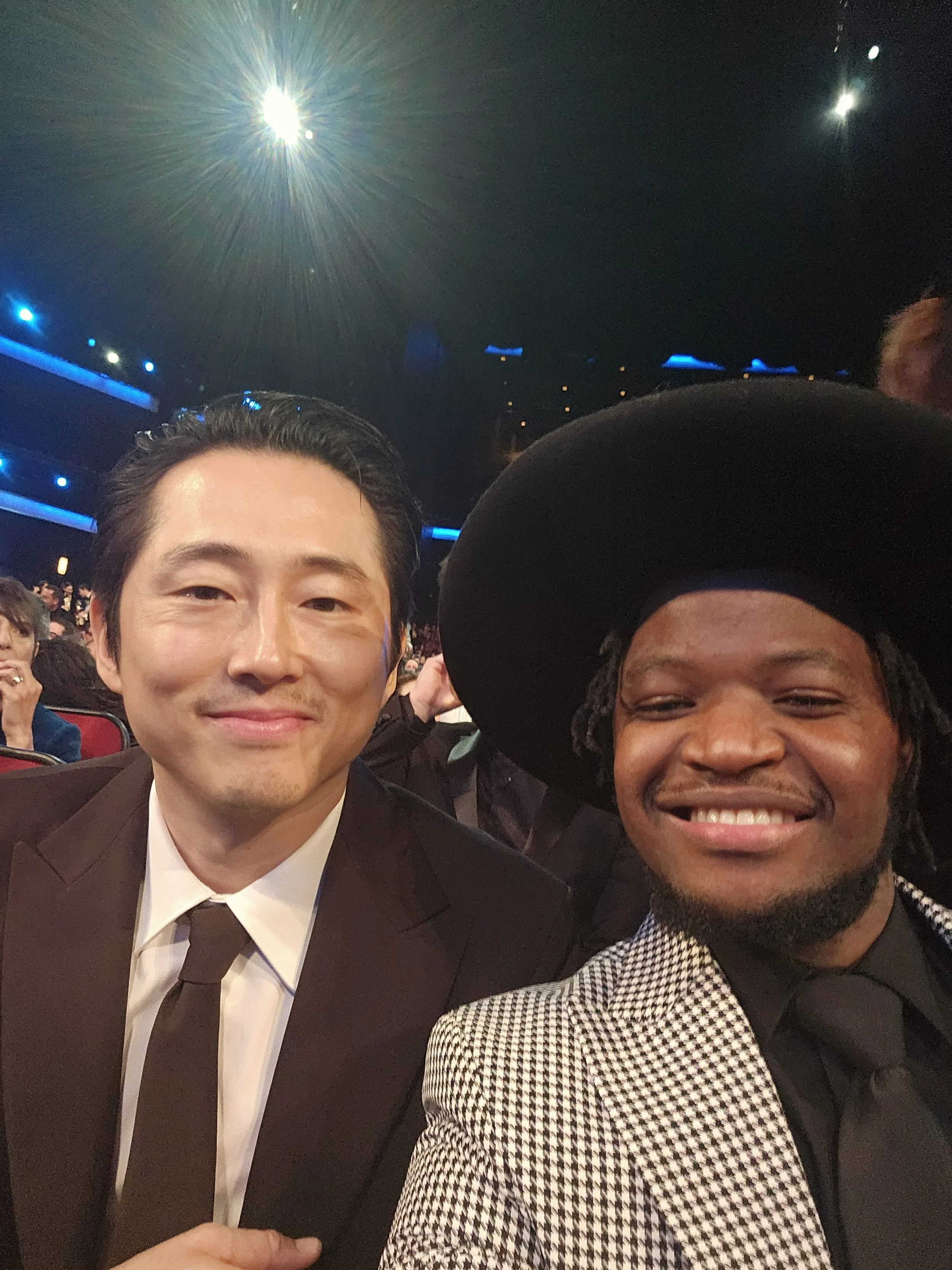 Two men smiling and taking a selfie at an event. One man is Asian, wearing a black suit, white shirt, and brown tie. The other man is Black, wearing a large black hat and a checkered blazer. The background shows a dark setting with blue and white lig