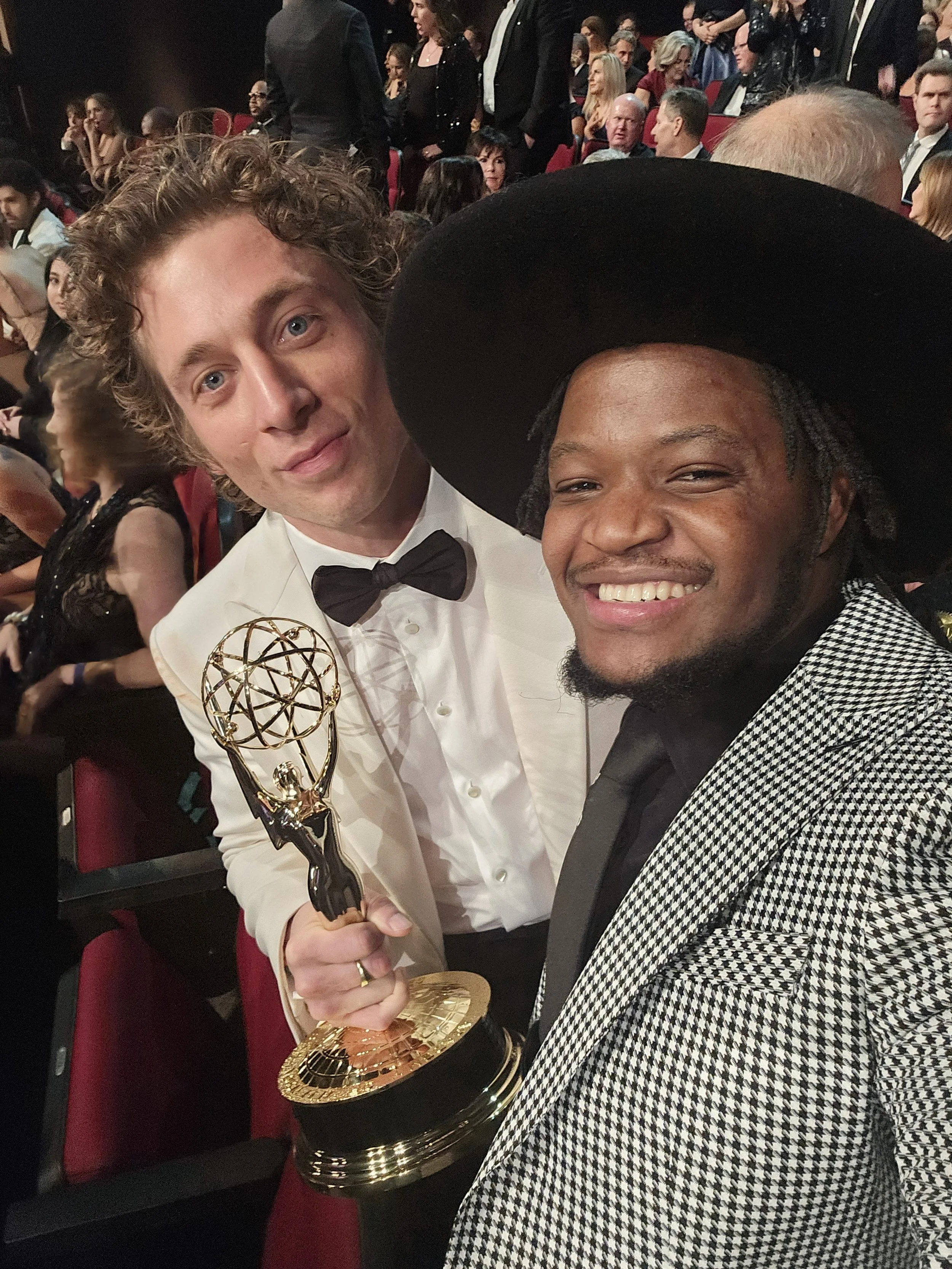 Two men at an awards ceremony, one holding an Emmy trophy, surrounded by an audience in formal attire.