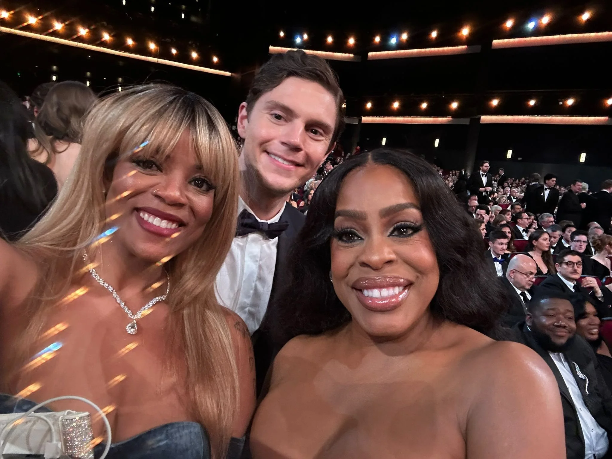Three smiling people taking a selfie at a formal event with many seated attendees in the background.