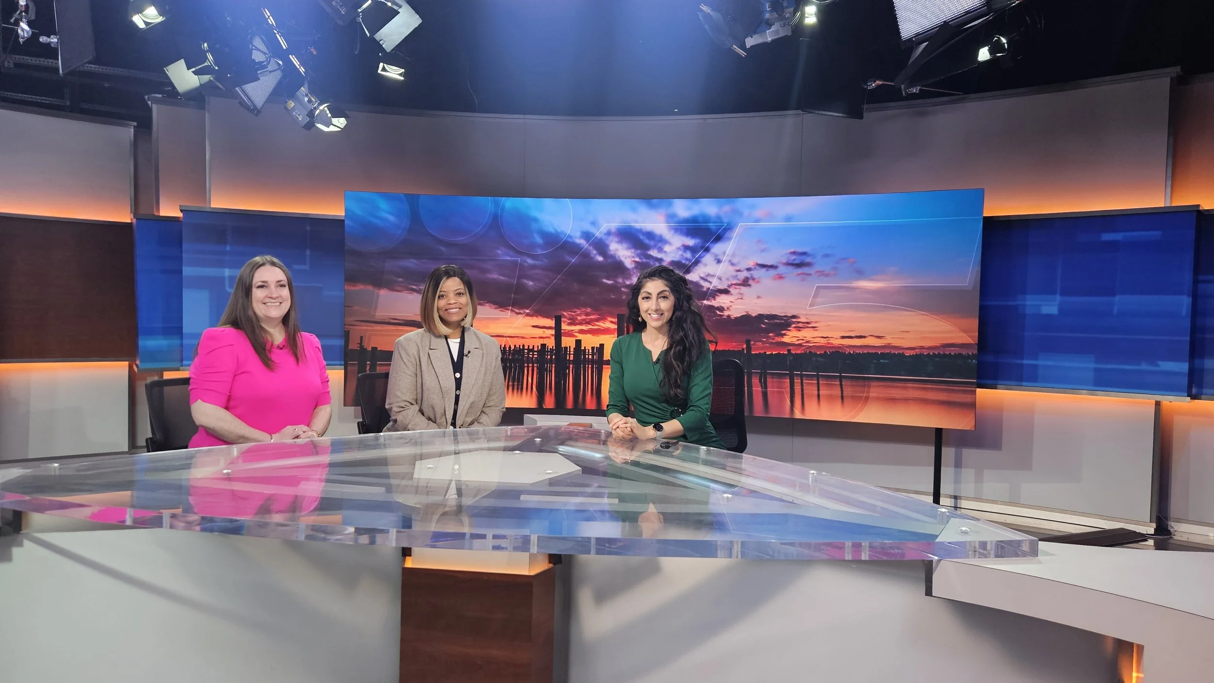 Three women sitting at a news desk in a TV studio with large screen monitors displaying a sunset over a body of water in the background.