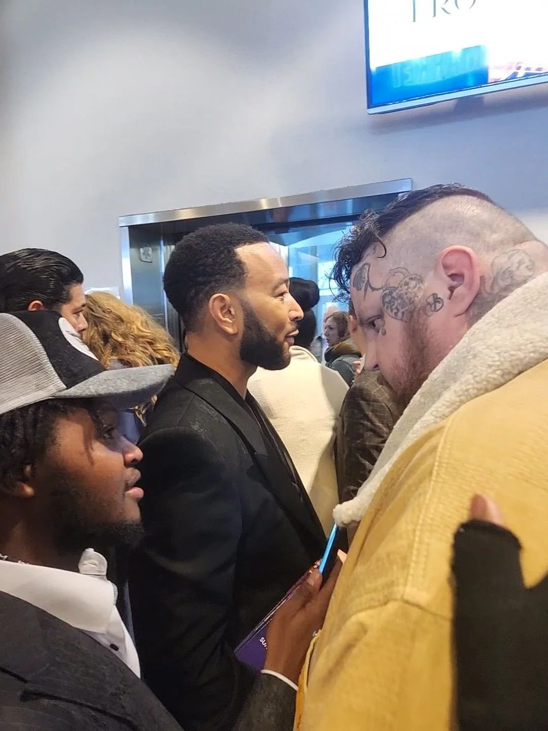 Crowded indoor scene with diverse people standing in line or gathering, some engaged with their phones, and a television on the wall showing an unclear image or text.