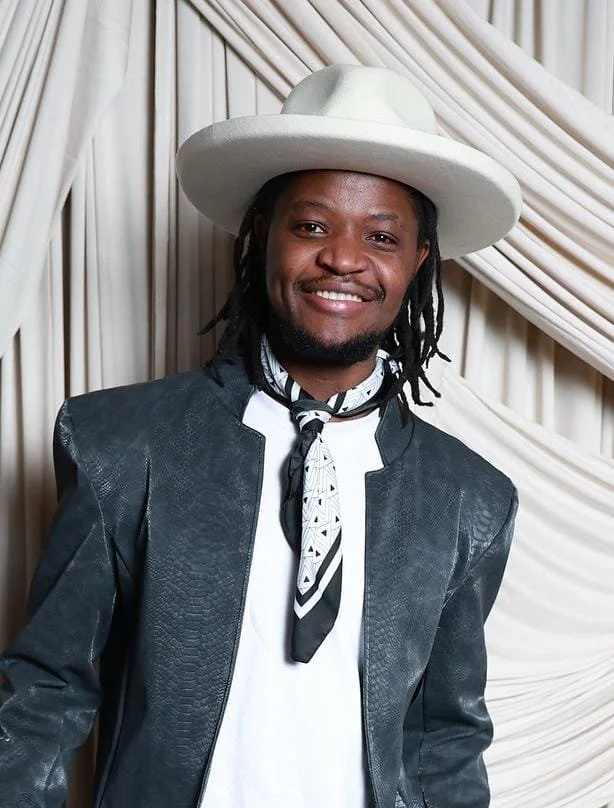 Smiling man with dreadlocks wearing a white hat, black leather jacket, and a black and white patterned neck scarf, standing in front of beige draped curtains.