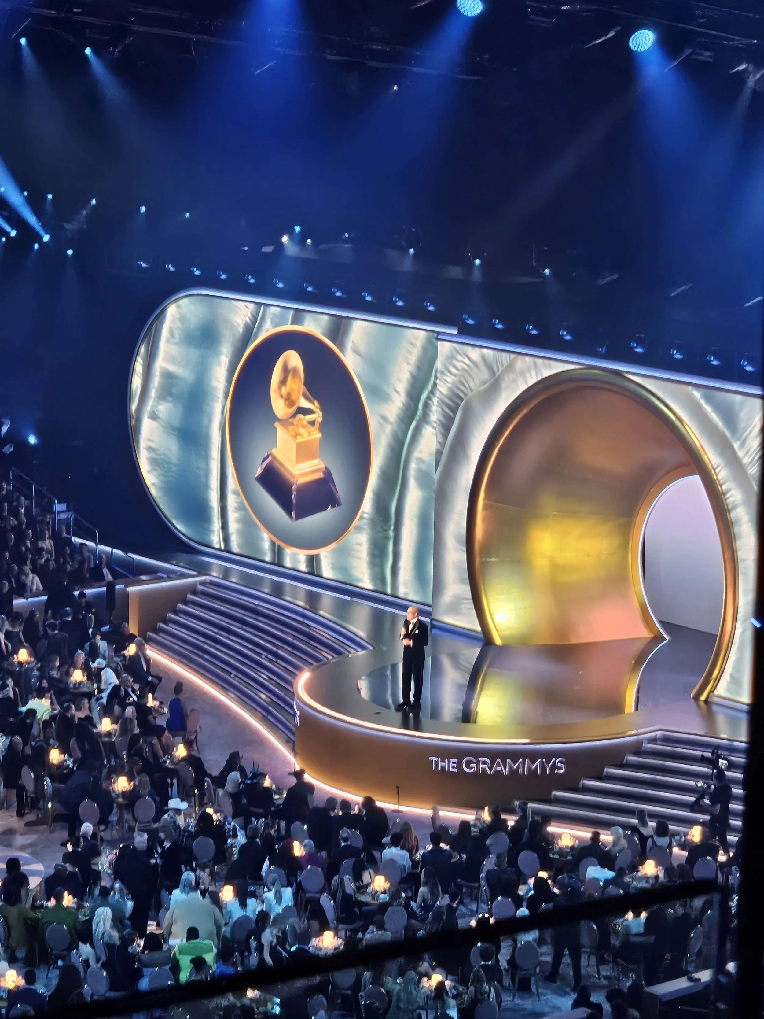 Stage at the Grammy Awards with large screen displaying a gold gramophone award, audience seated at tables with lit candles, and a presenter on stage.