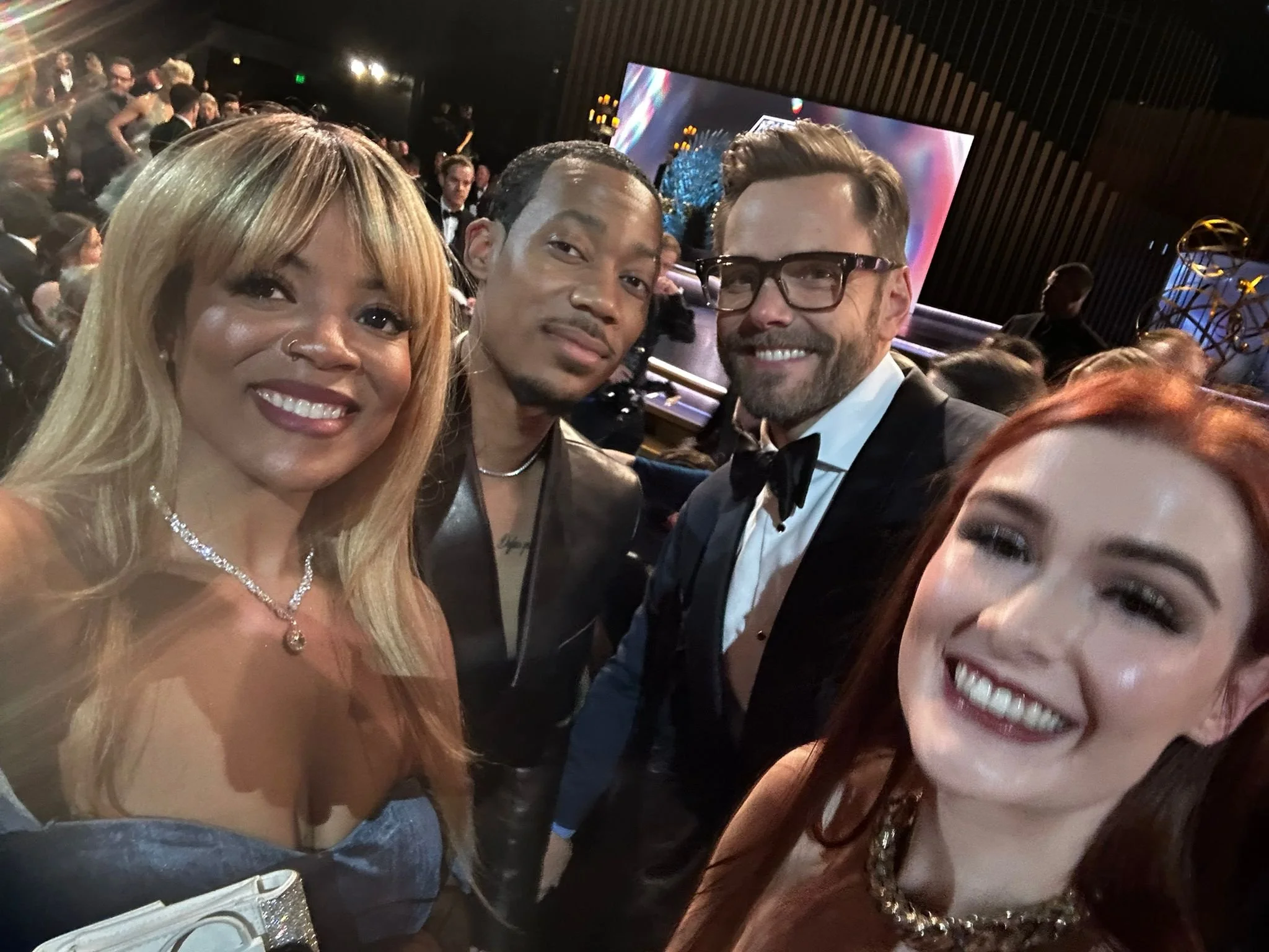 Four people dressed in formal attire smiling at a party or event with a large screen and other guests in the background.