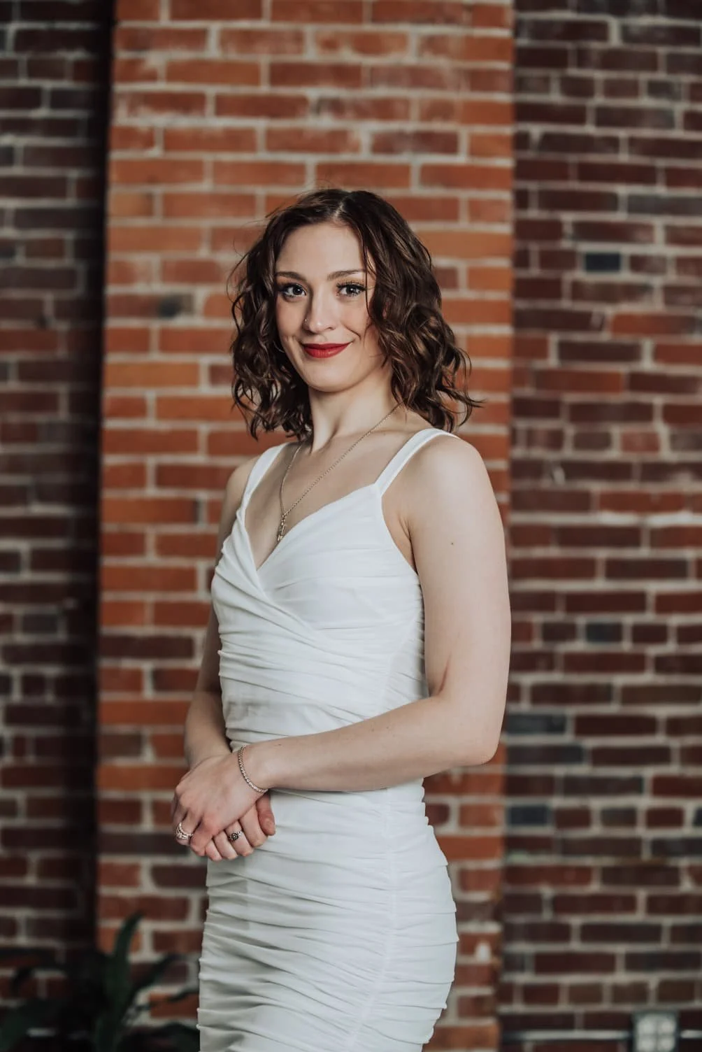 A young woman with curly brown hair and red lipstick is standing in front of a brick wall. She is wearing a white, ruched sleeveless dress and jewelry, smiling at the camera.