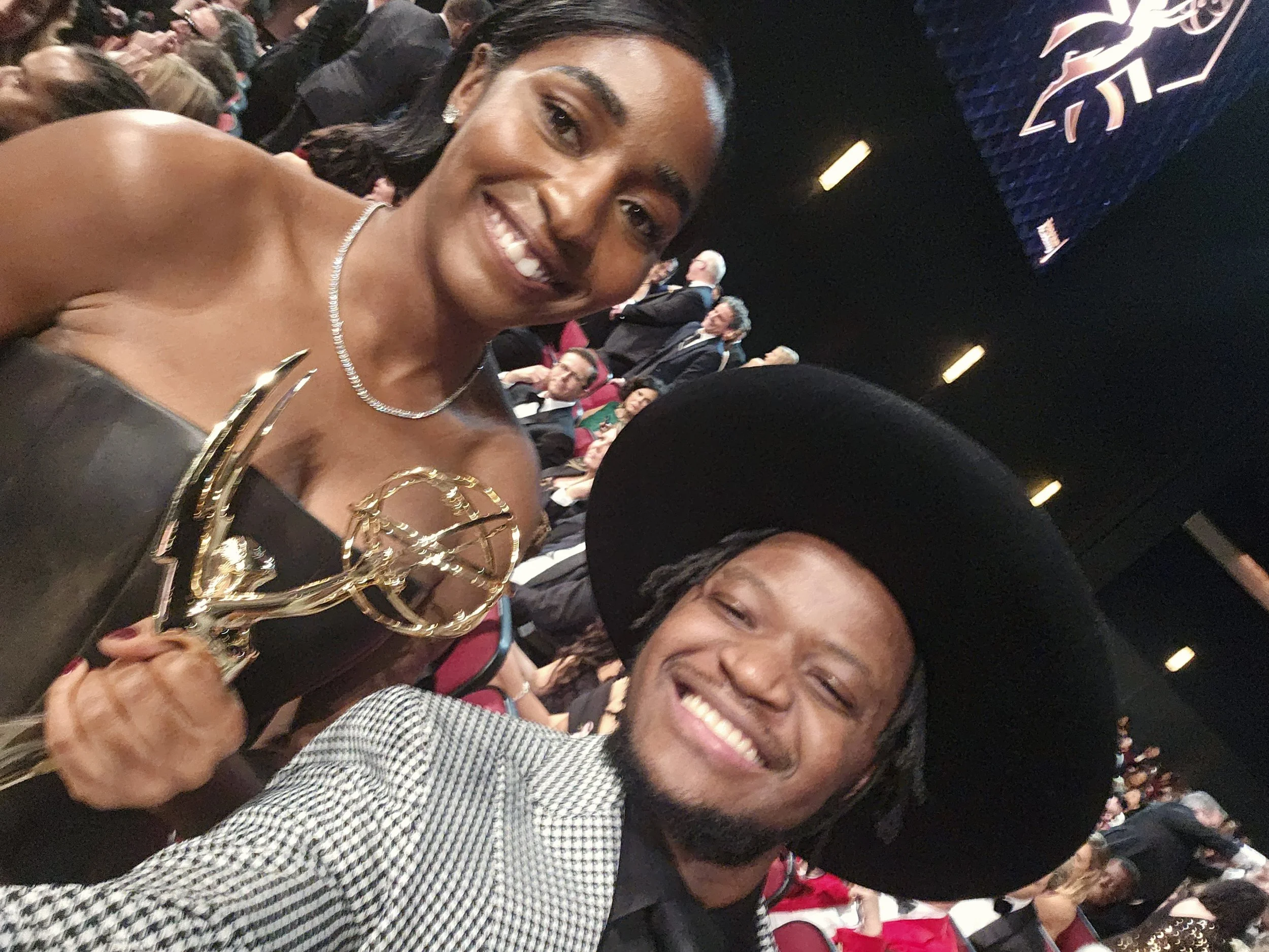 A woman smiling and holding an award, standing next to a man smiling and taking a selfie, in an audience setting.