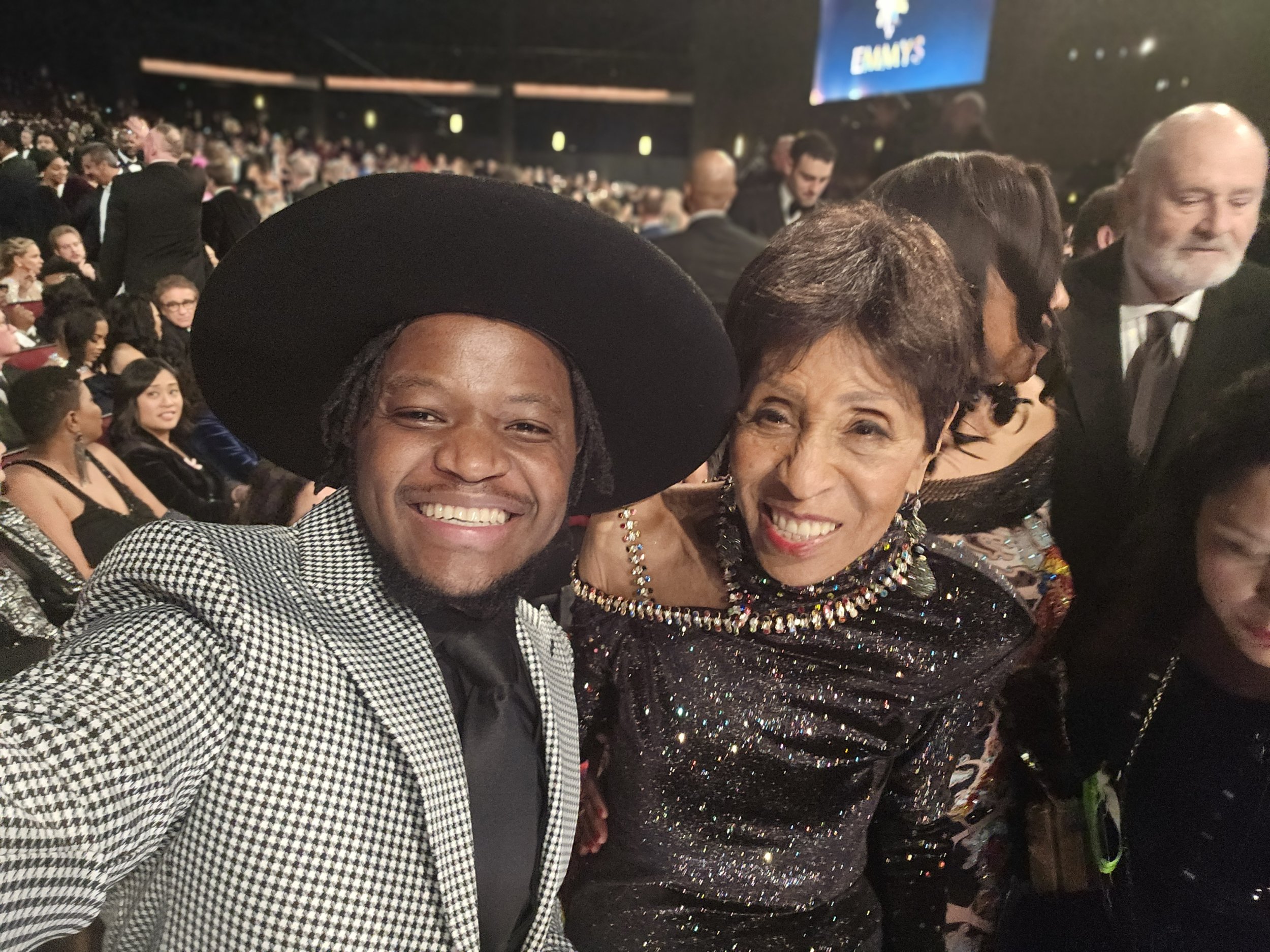 People at an award ceremony or event, dressed in elegant attire, seated in a large hall with a stage and a screen in the background.