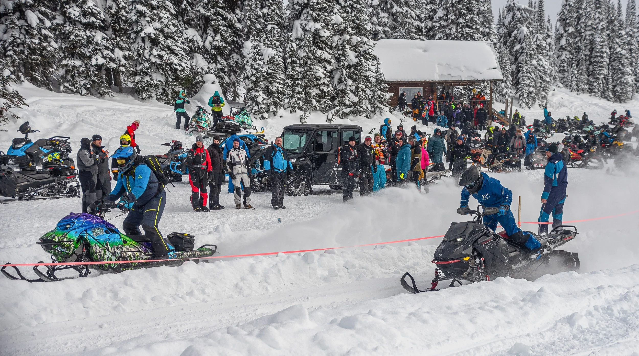 Snow-covered outdoor scene at a winter event with numerous people, snowmobiles, and a small wooden building in the background surrounded by trees.