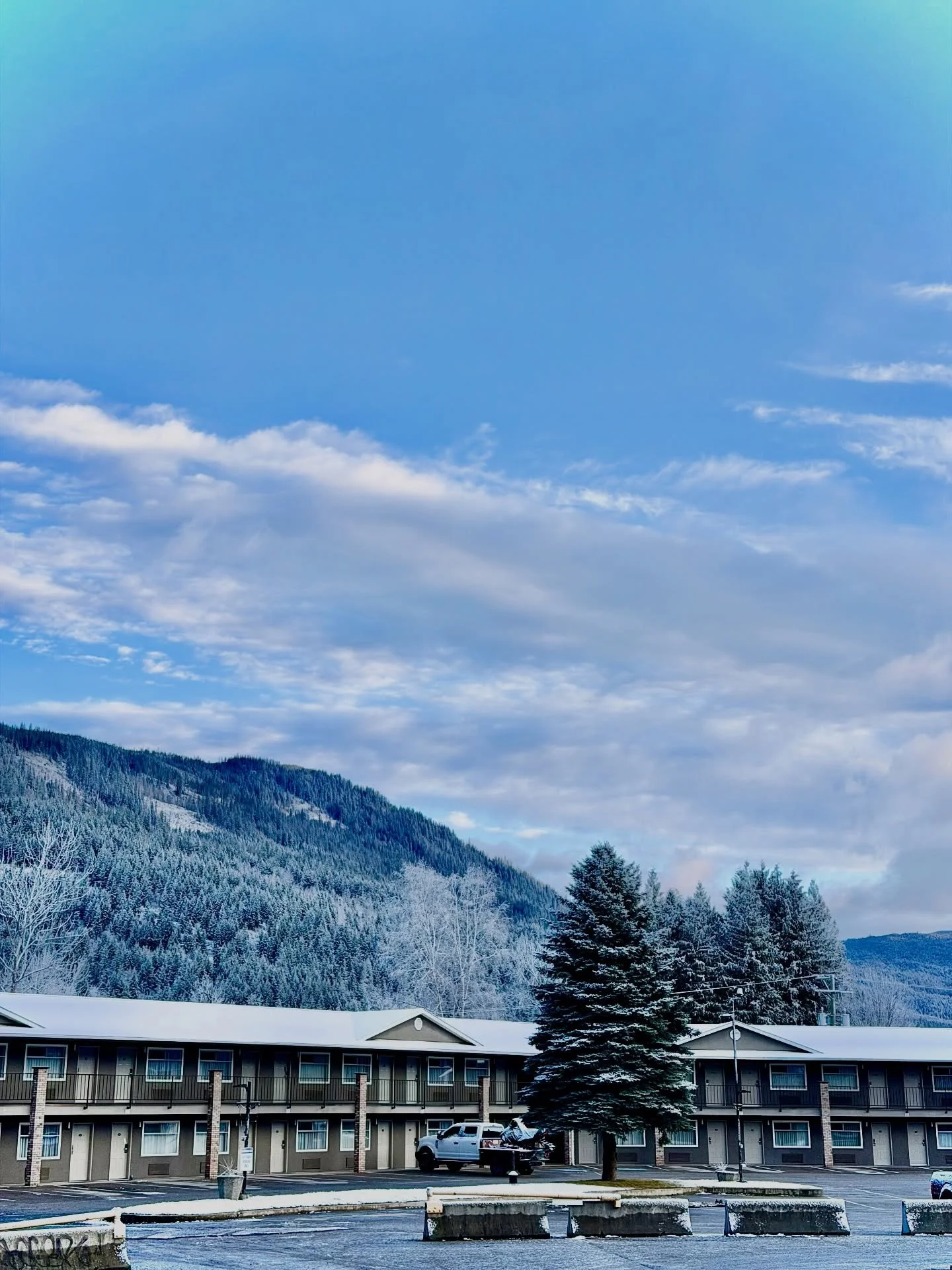 A parking lot in front of a low-rise building with a mountain range covered in snow in the background, and a partly cloudy sky above.