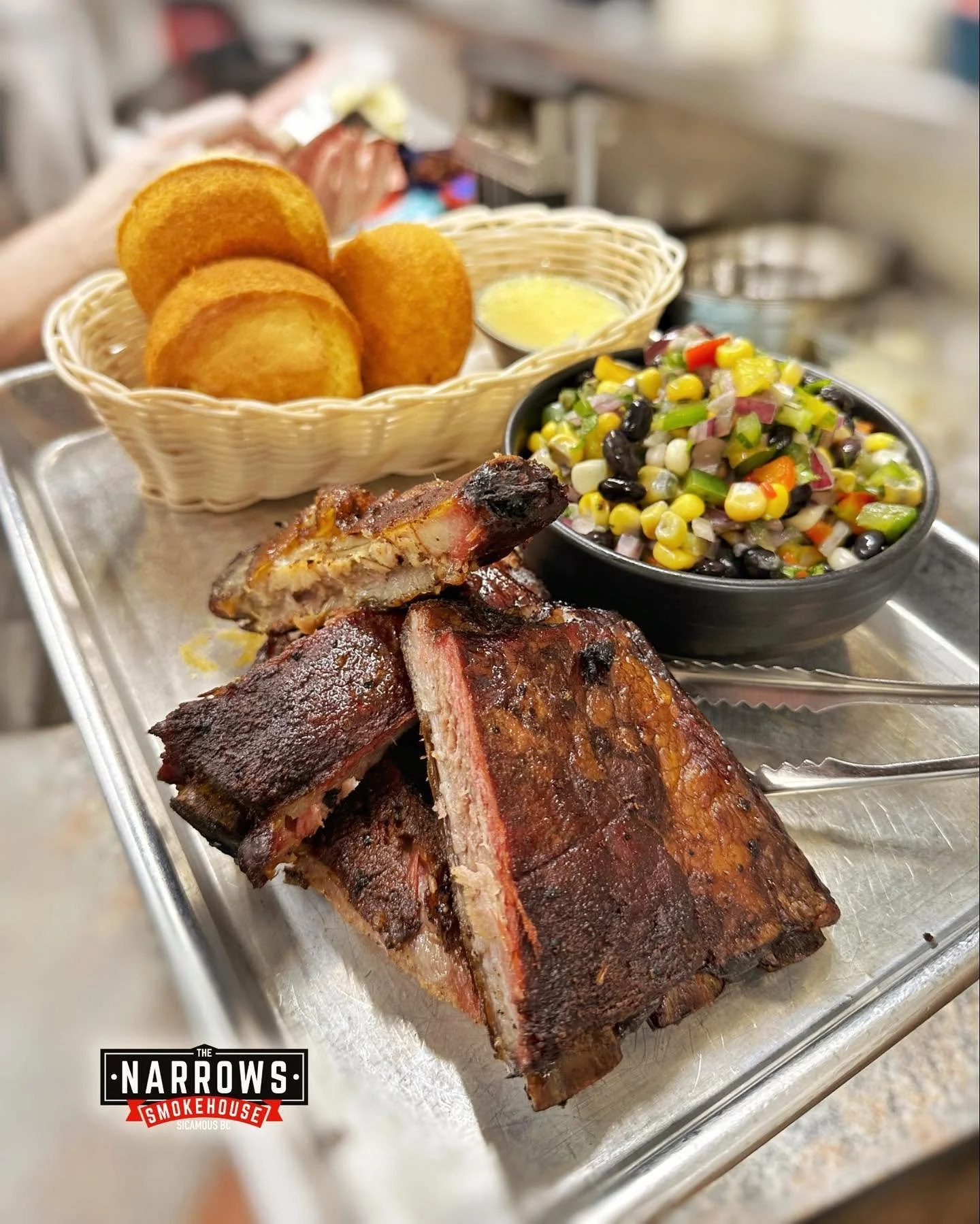 Barbecue ribs with a dark, crispy exterior and juicy interior on a metal tray, accompanied by cornbread muffins in a basket, a bowl of colorful corn salad with vegetables, and a small dish of melted butter, at The Narrows Smokehouse.