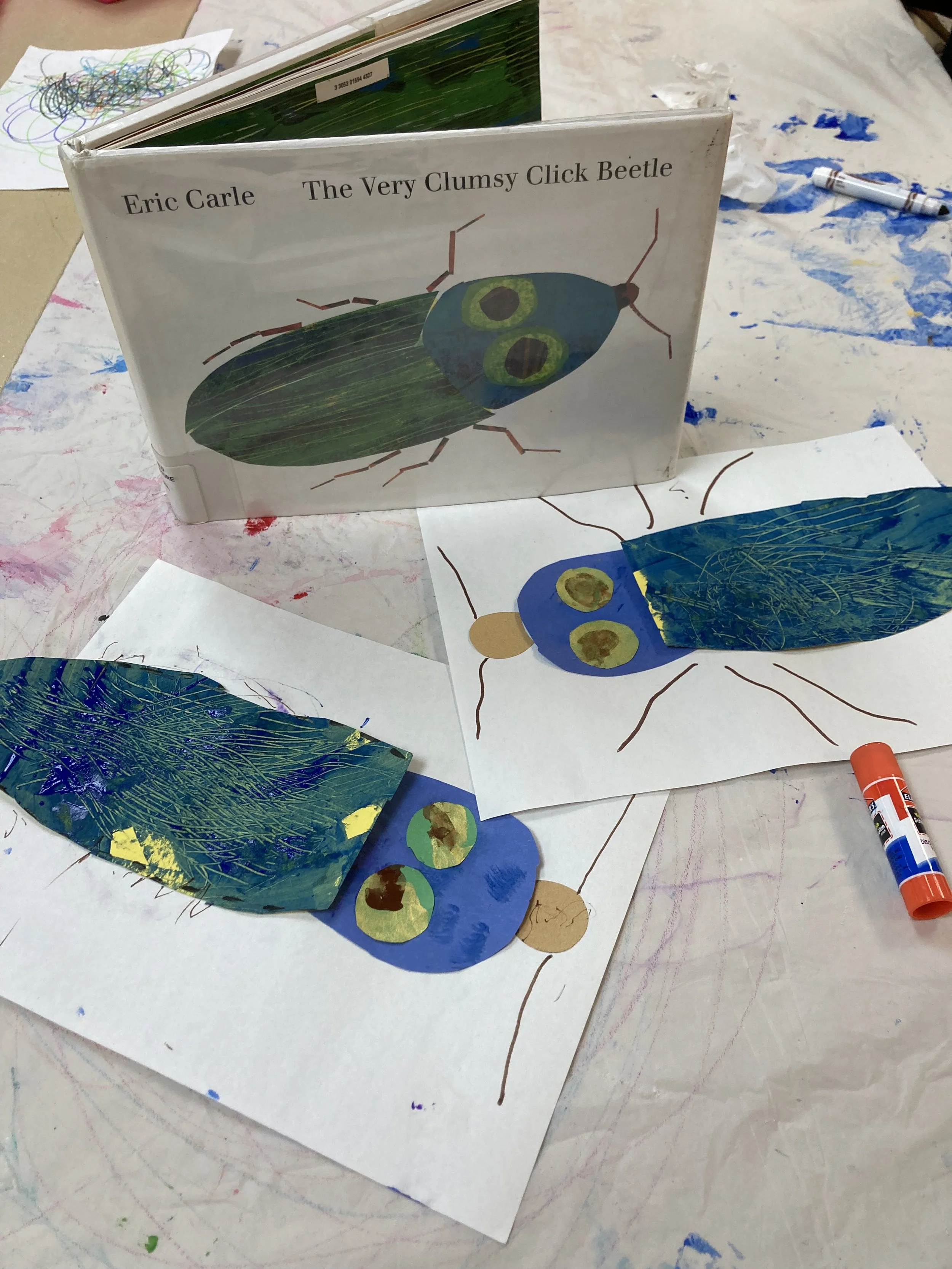 Craft project featuring a paper beetle inspired by Eric Carle's "The Very Clumsy Click Beetle," with painted and glued paper wings and drawn body parts on sheets of paper, surrounded by art supplies and colorful paint marks.