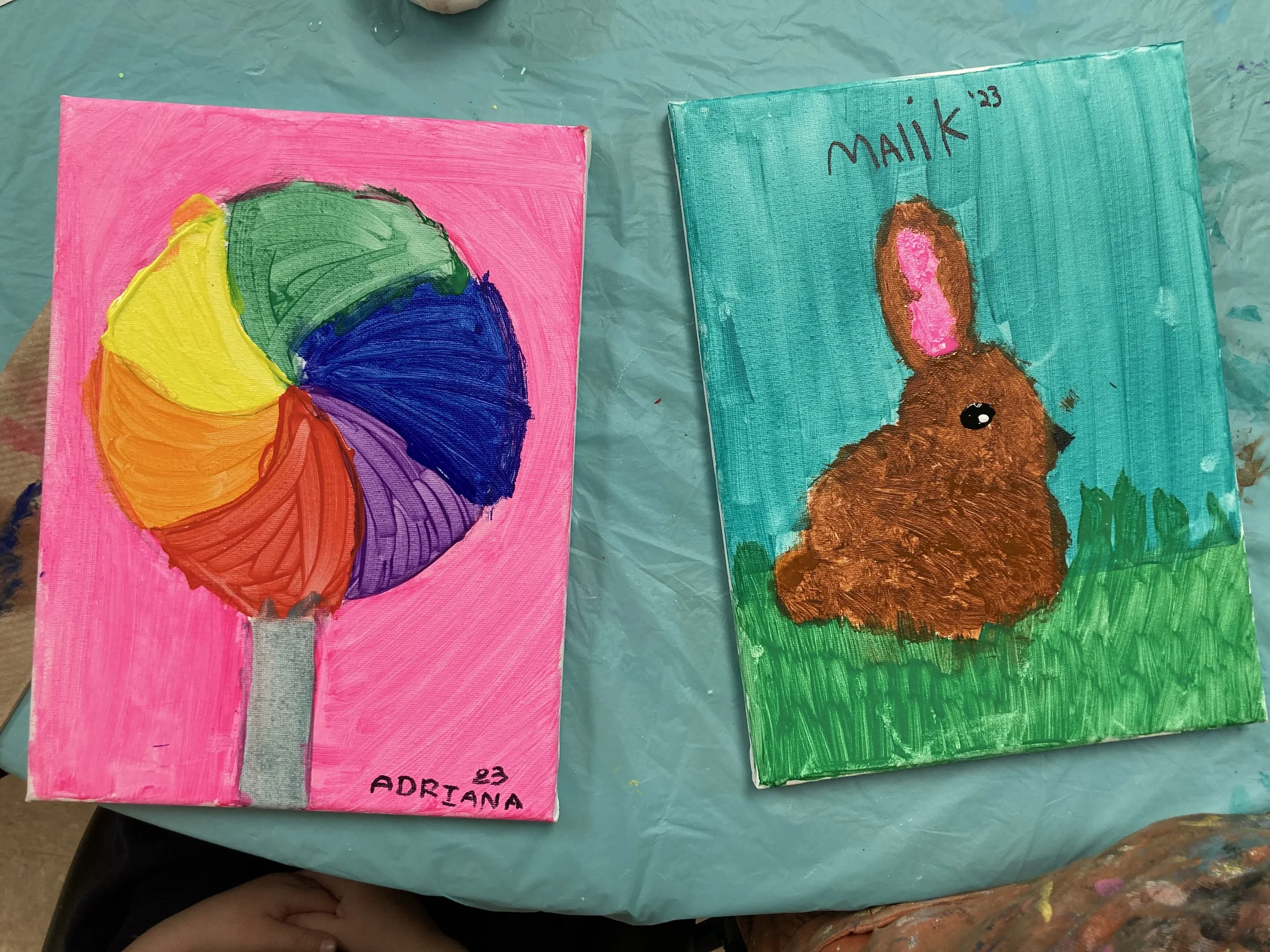 Two colorful paintings on canvas, one of a rainbow-colored ice cream on a pink background, and the other of a brown bunny with pink inner ears and a black eye on a green grassy background.