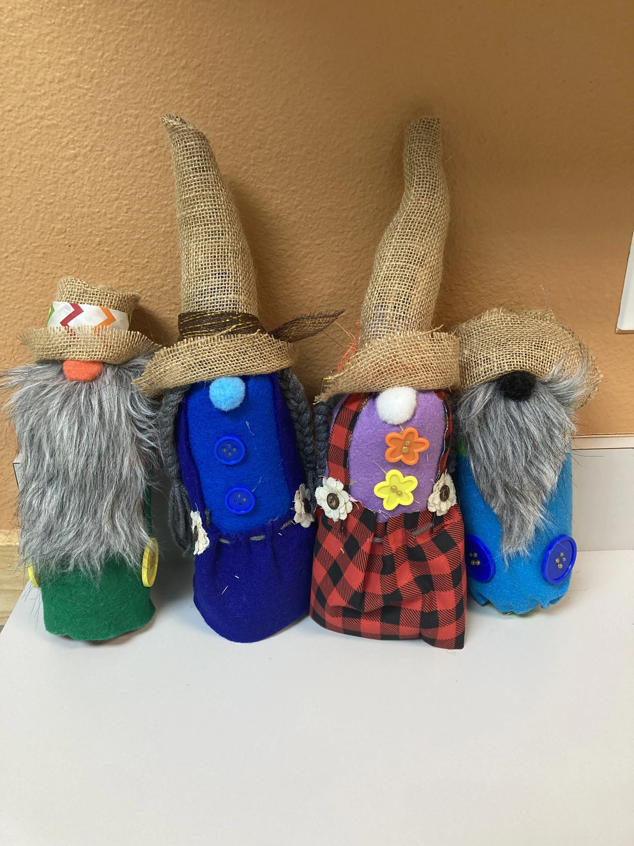Four handmade gnome dolls with tall, jute hats, yarn beards, and colorful clothing, standing on a white surface against a tan wall.