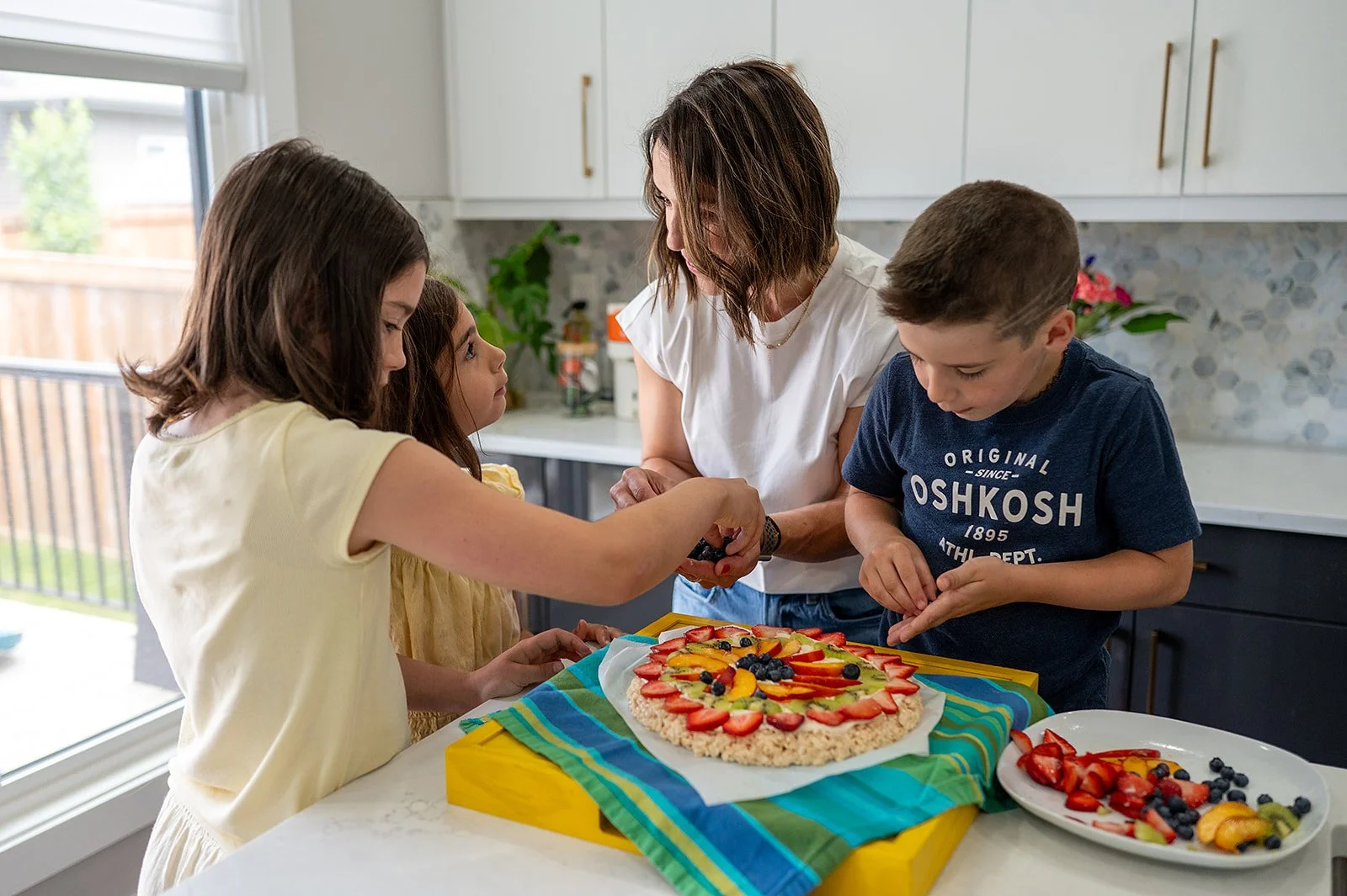 Megan Wallace and her three children decorate a fruit pizza together in a bright family kitchen.