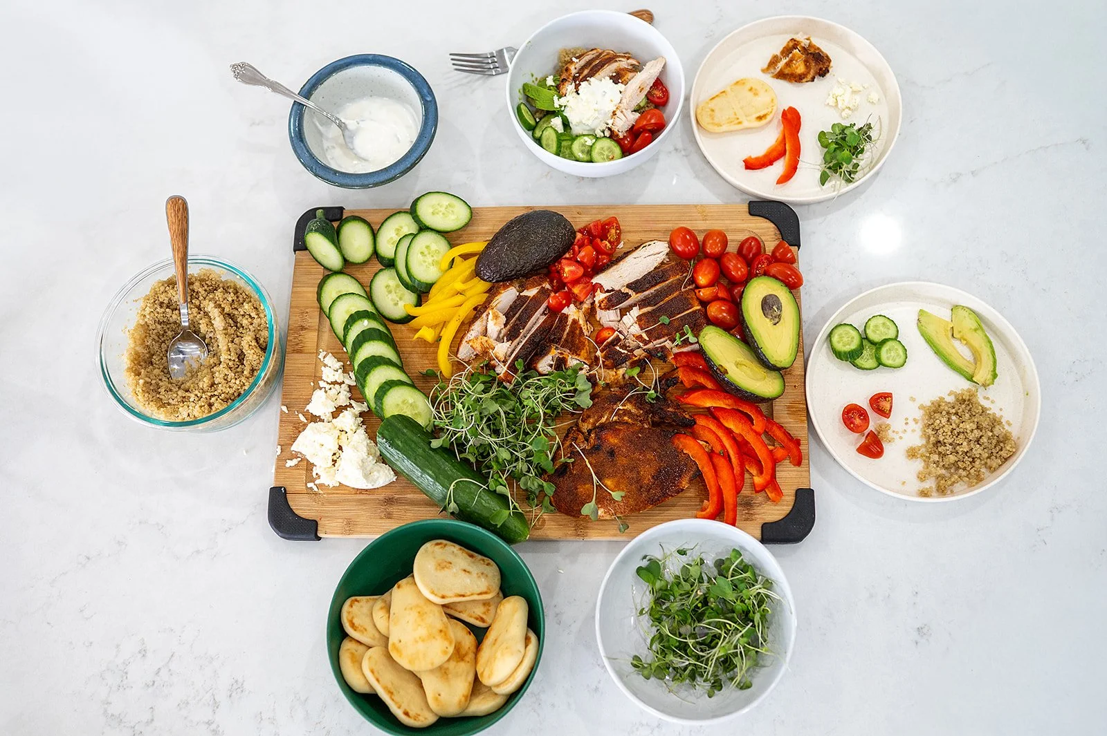Wide flat-lay spread of a chicken quinoa bowl bar with pita bread, tzatziki, and fresh vegetables