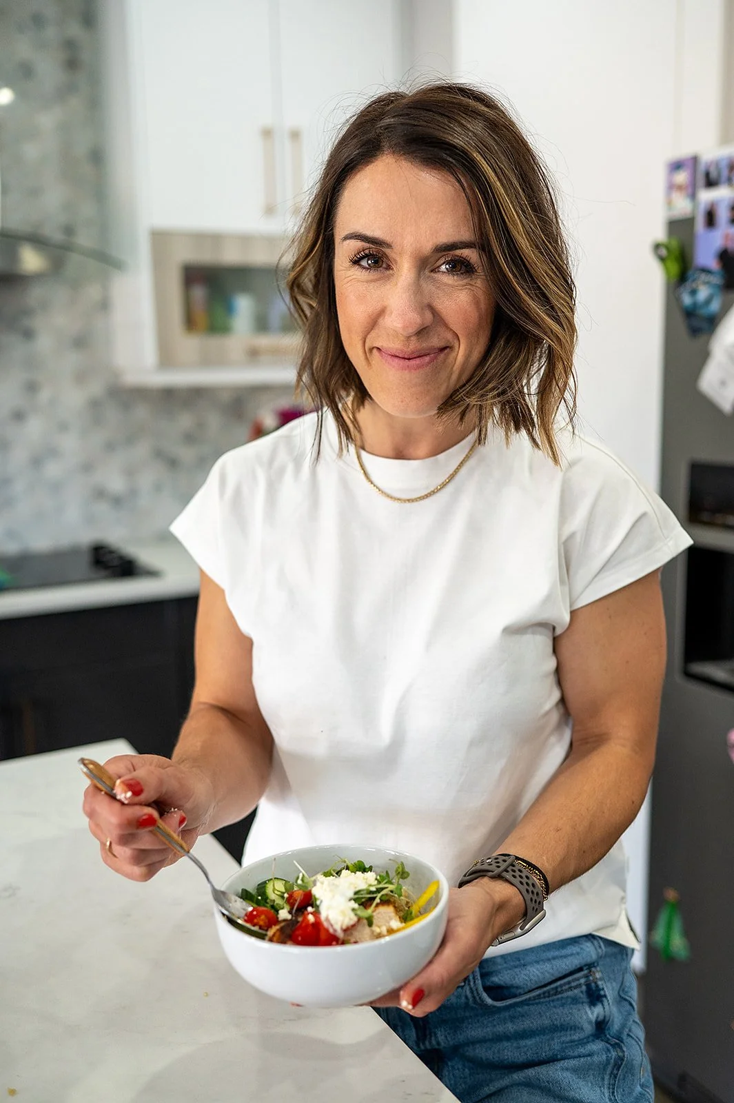 Registered dietitian smiling while holding a colorful chicken, quinoa, and vegetable bowl in her kitchen.
