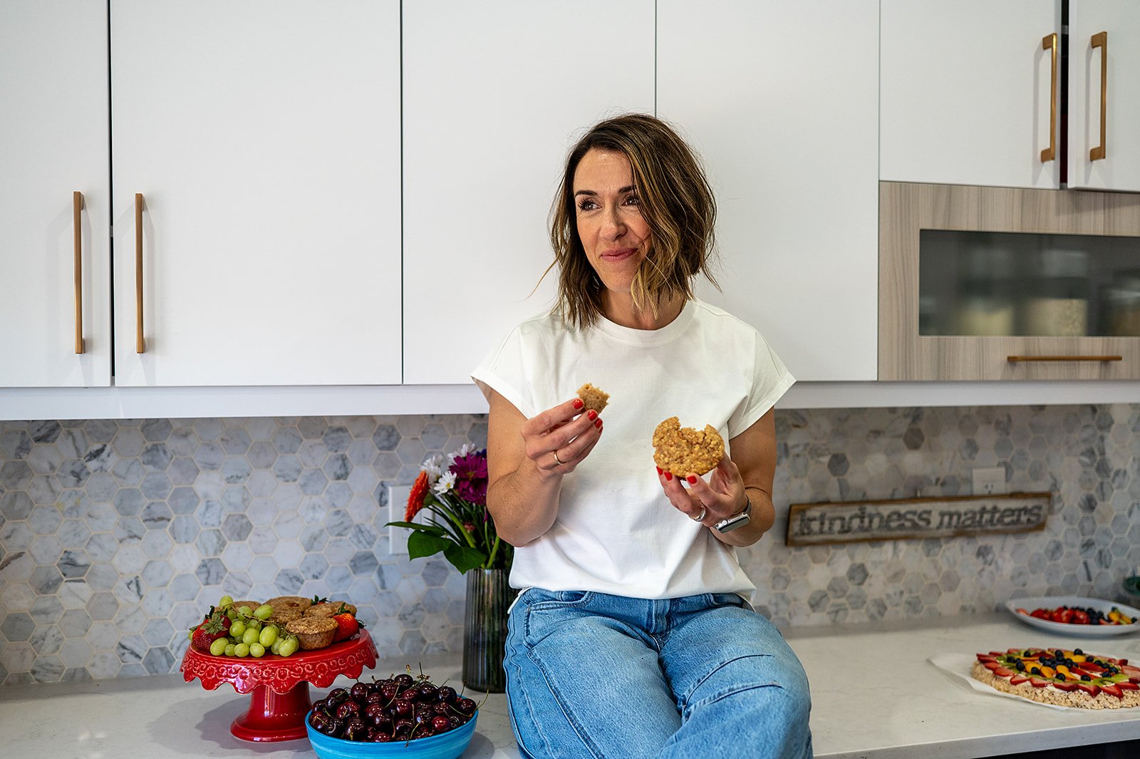 Registered dietitian enjoys a homemade oat muffin surrounded by fresh fruit in her kitchen.