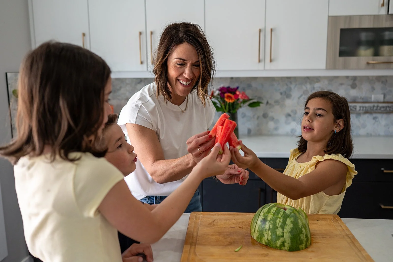 Megan Wallace hands freshly cut watermelon spears to her children gathered around the kitchen counter.