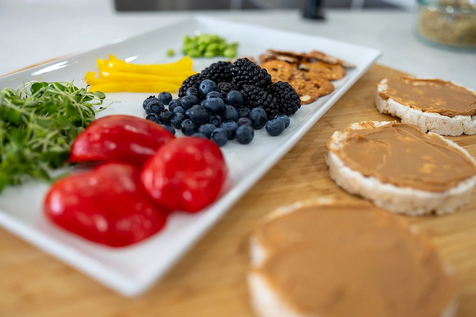 Family-style snack platter with mini peppers, blueberries, blackberries, pretzels, and peanut butter rice cakes.