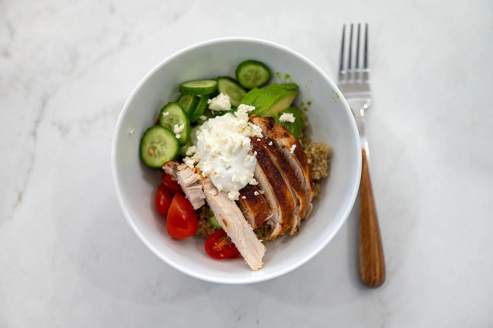 Overhead shot of a chicken quinoa bowl topped with feta, tzatziki, avocado, and tomatoes