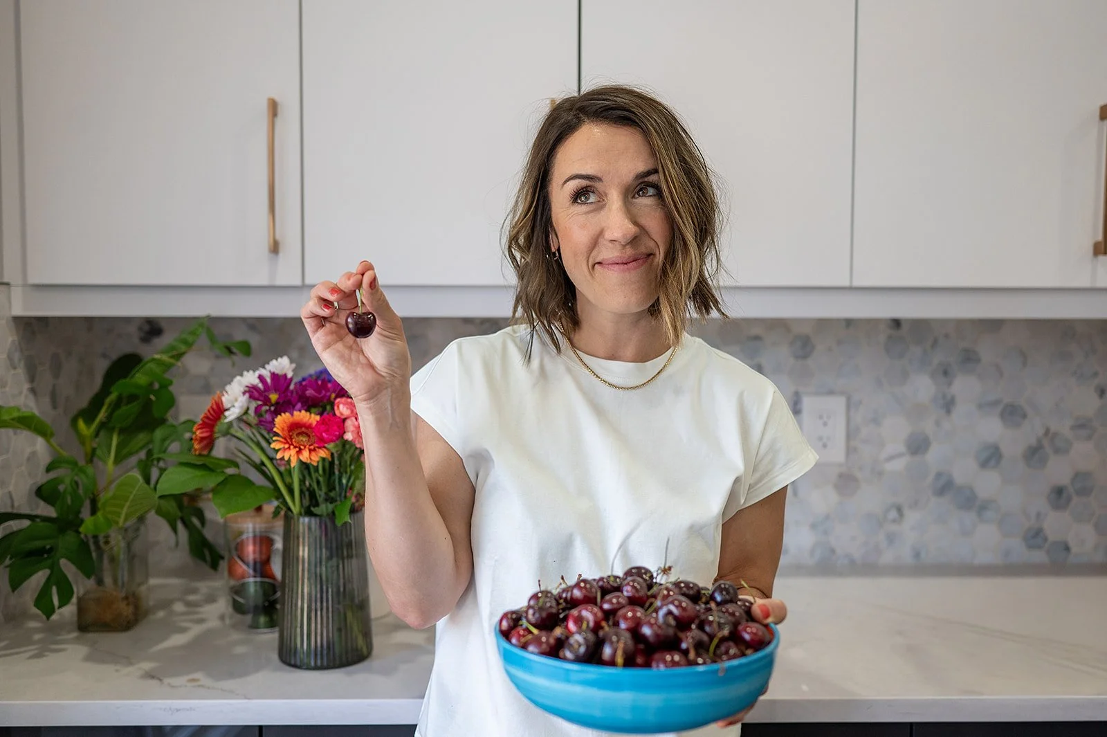 Megan Wallace in a white kitchen holding up a cherry, smiling next to a blue bowl of fresh cherries and flowers.