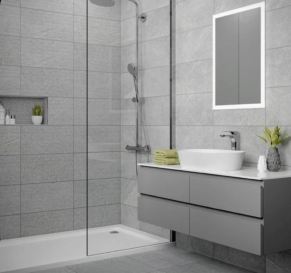 Modern bathroom with a walk-in shower, gray tiled walls, a gray vanity with a white vessel sink, a rectangular mirror, and decorative items like green towels, a plant, and small containers.