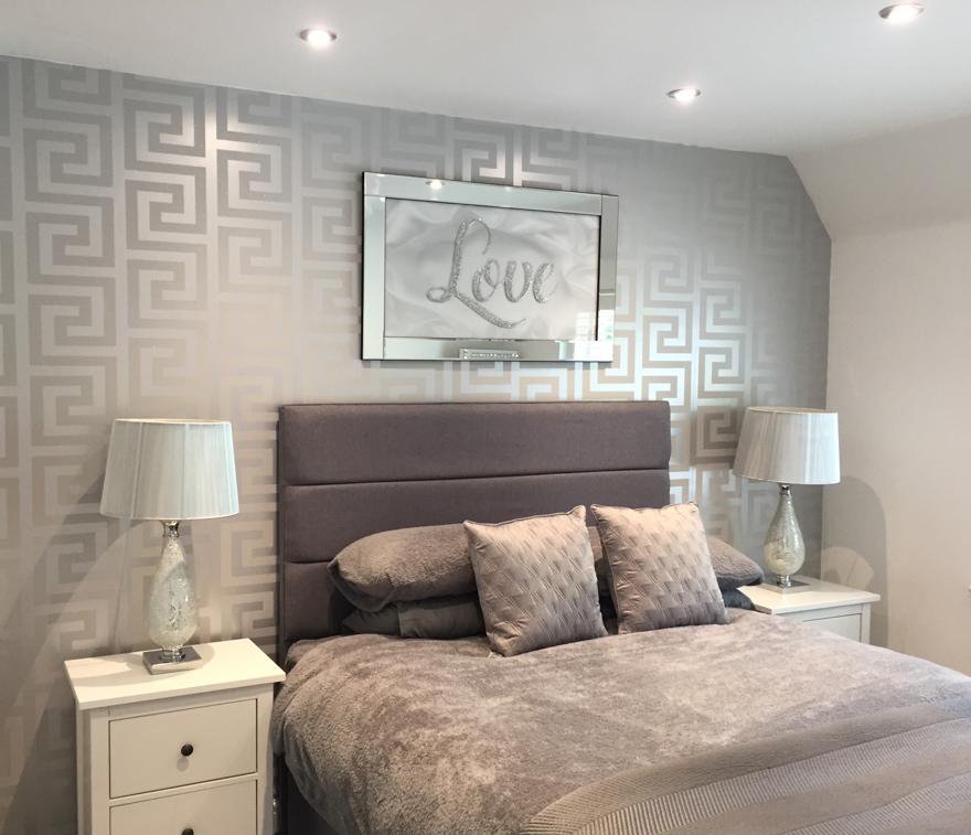Bedroom with gray patterned wallpaper, gray upholstered headboard, two white nightstands with glass lamps, and a framed decorative word 'Love' above the bed.