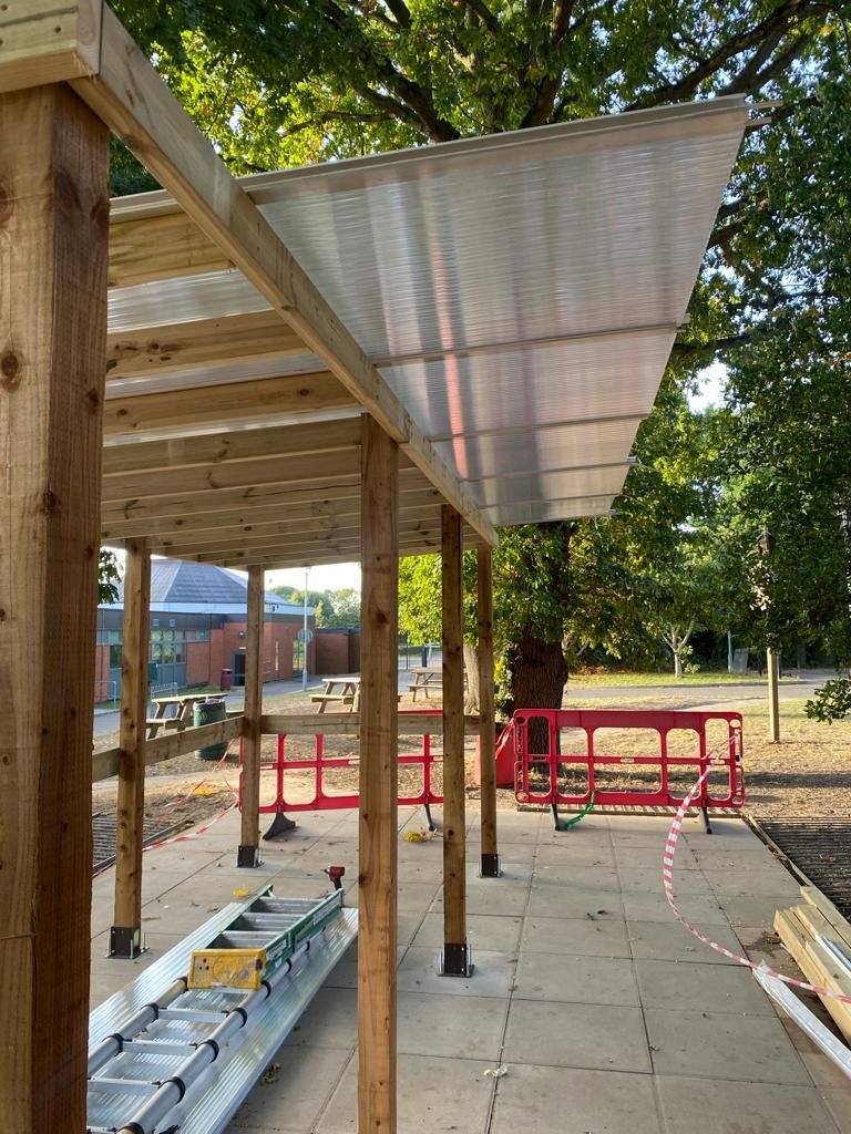 Construction of a wooden outdoor shelter with a metal roof in progress, surrounded by safety barriers and construction tools, trees in the background.