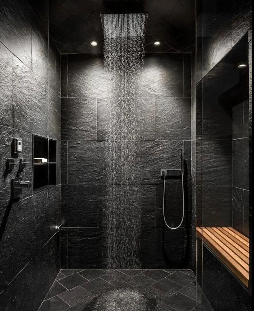 Modern shower with black tiled walls, a rain shower head, built-in shelf, handheld shower, and wooden bench.