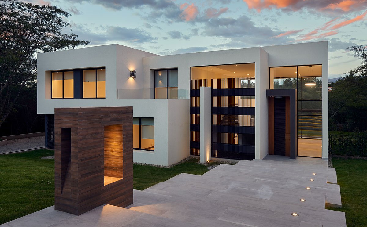 Modern two-story house with large glass windows and a wooden sculpture in front, set against a sunset sky.