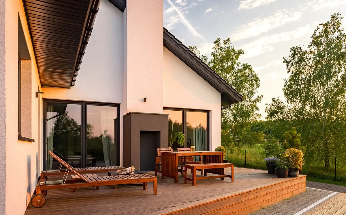 A wooden deck attached to a modern house with large glass doors, outdoor furniture, potted plants, and trees in the background during sunset.