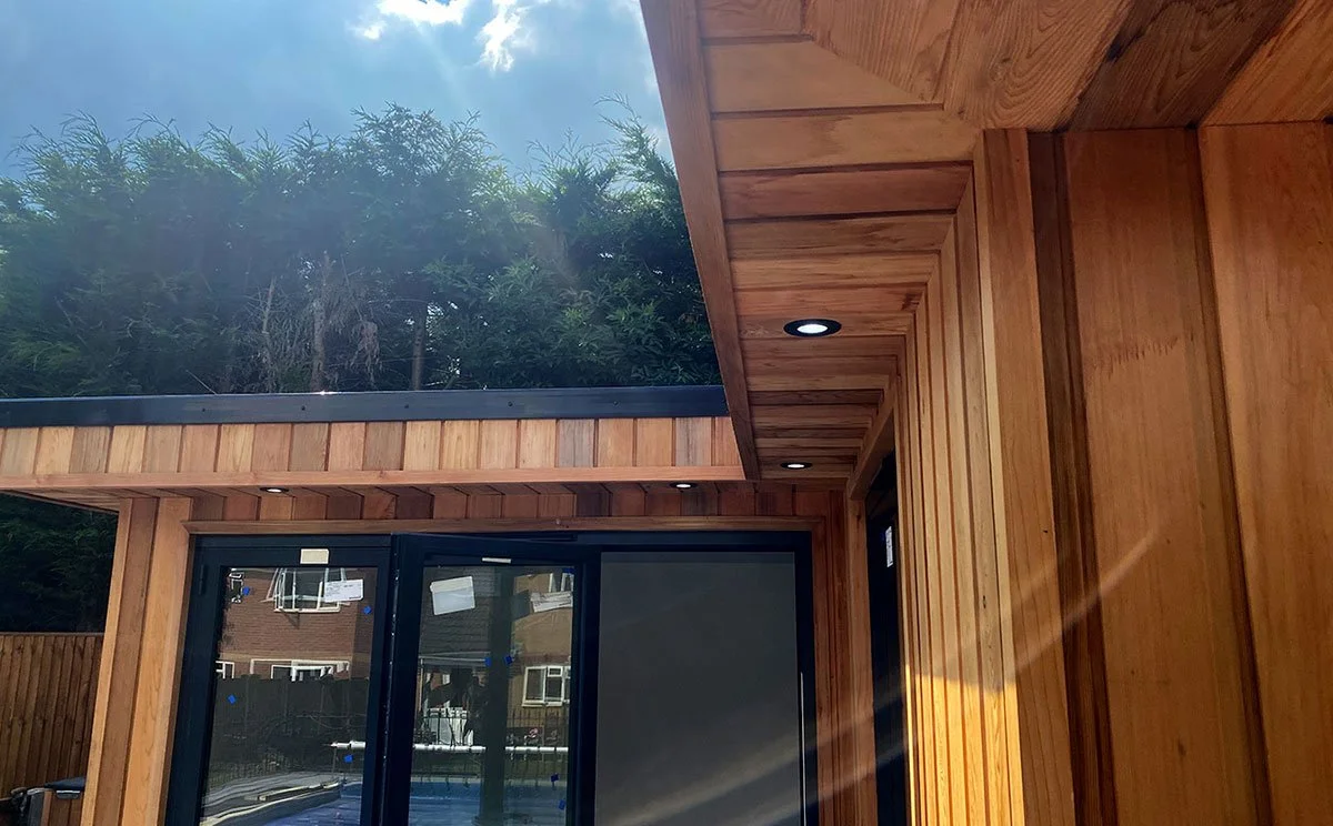 Close-up of a modern house exterior with wooden siding, ceiling lights, and a sliding glass door reflecting surrounding houses and trees under a cloudy sky.