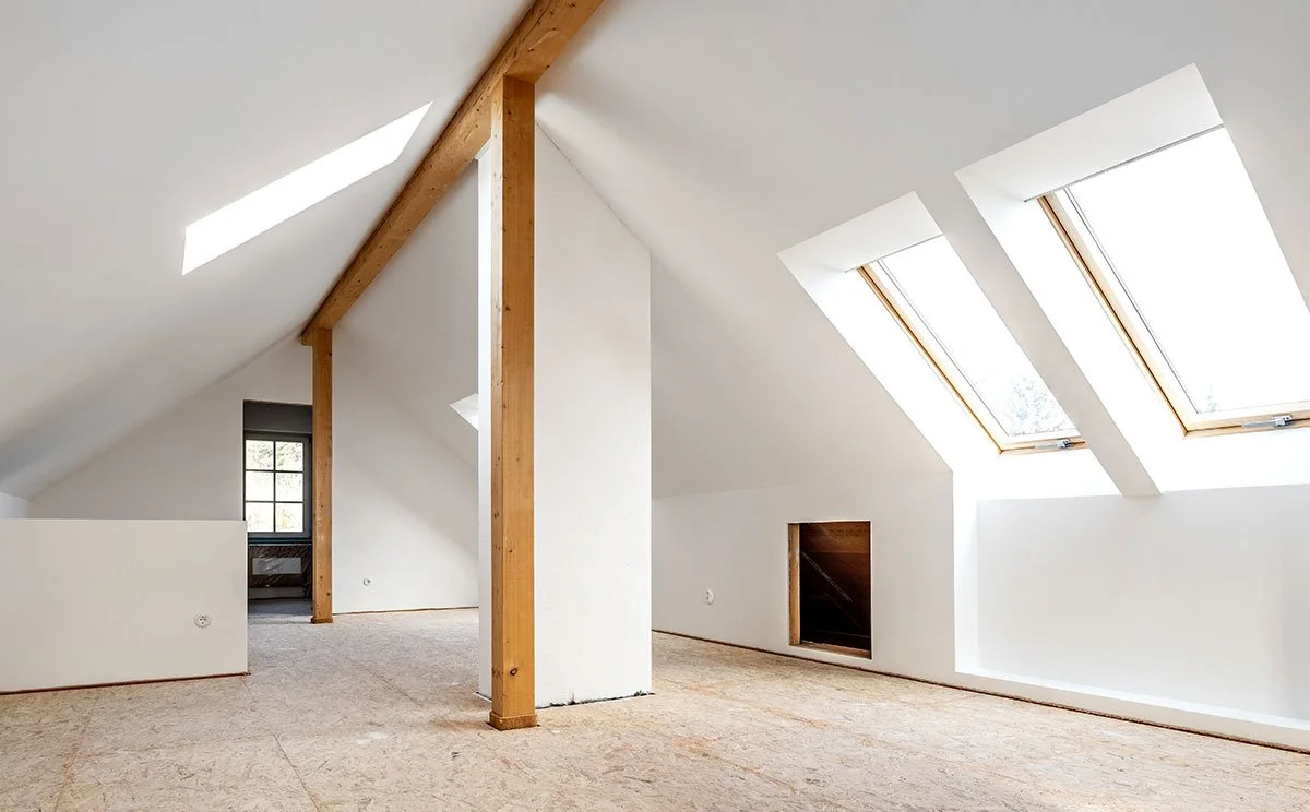 Empty attic room with skylight windows and wooden beams, unfinished floors, and white walls.