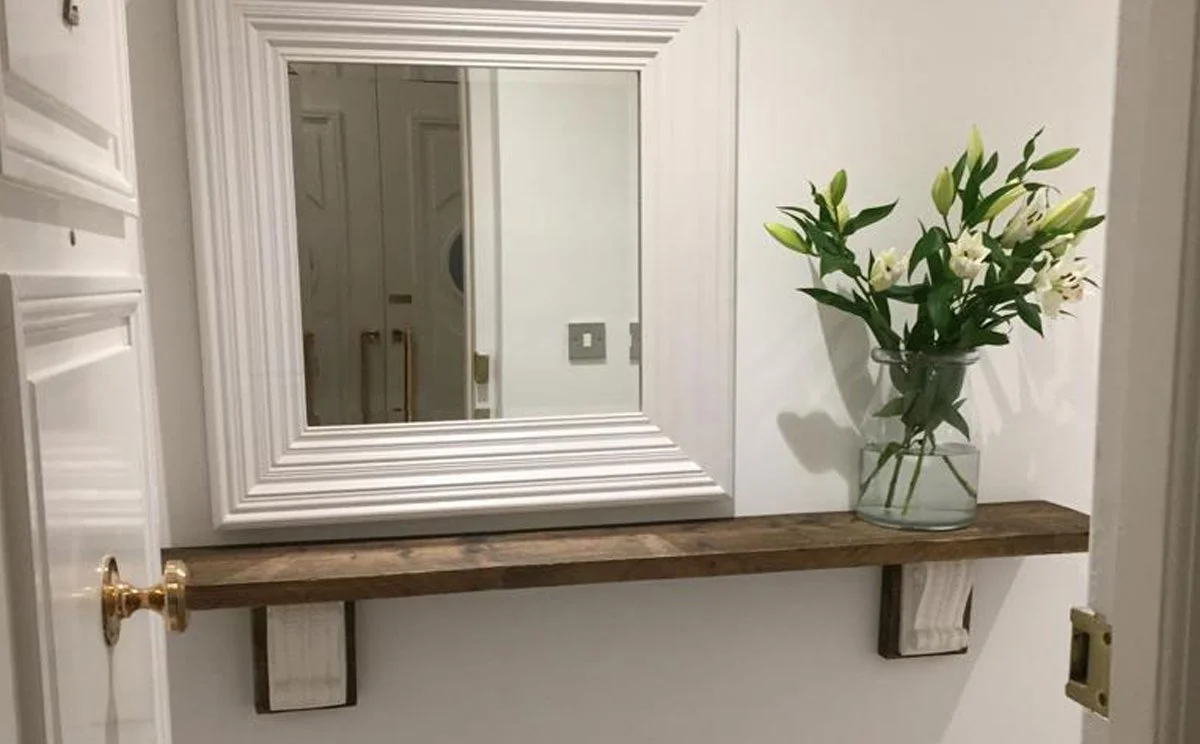 a white-framed mirror hanging on a white wall above a rustic wooden shelf with a glass vase filled with white lilies and greenery