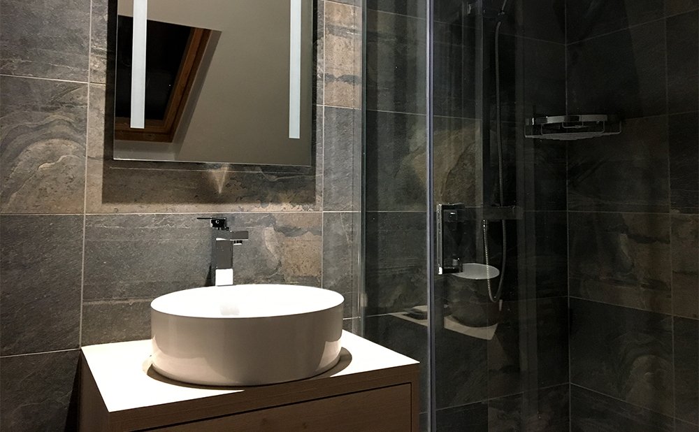 Modern bathroom with dark stone tiles, a white oval vessel sink on a wooden countertop, a rectangular mirror, and a glass-enclosed shower area.