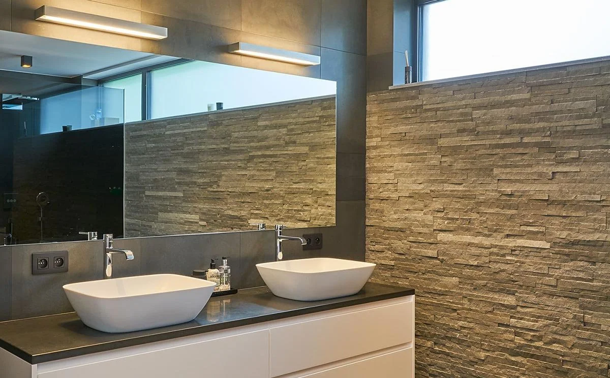 Modern bathroom with two vessel sinks on a dark countertop, large mirror, wood-textured wall, bathroom soap dispensers and lotion bottles, and a window with natural light.