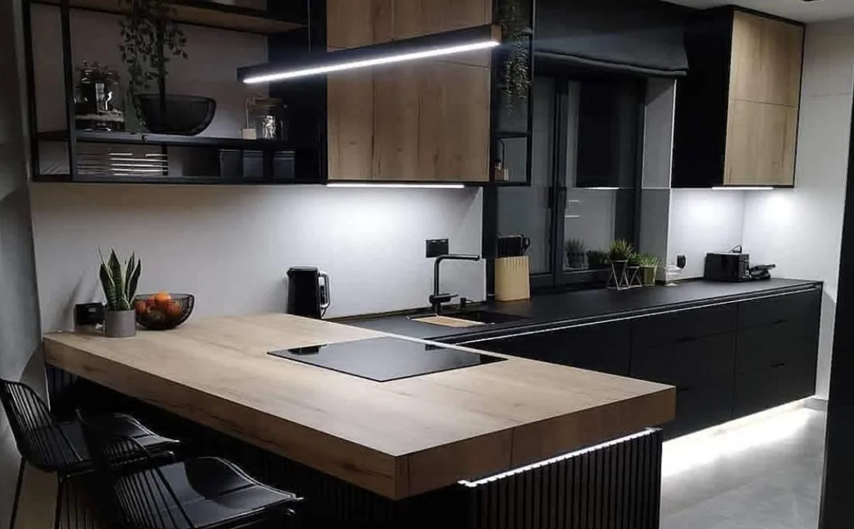 Modern kitchen with black and wood cabinets, a large light-colored wooden island with a black induction cooktop, black chairs, and minimal decor including plants and small appliances. Overhead lighting provides illumination.