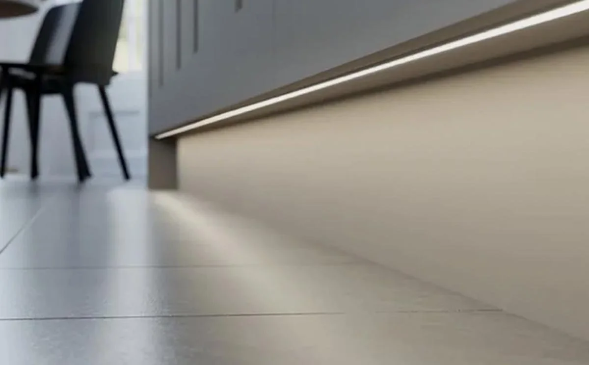 Close-up view of the bottom of a kitchen cabinet with under-cabinet lighting, kitchen floor, and part of a dining area with chairs.