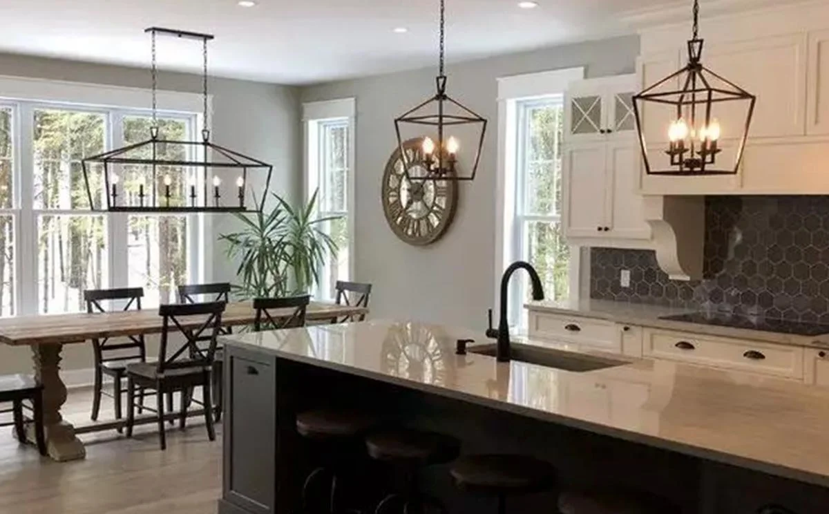 Kitchen and dining area with large windows, two black chandeliers, a wooden dining table with six chairs, a kitchen island with a sink, white cabinets, a decorative clock on the wall, and a plant by the window.