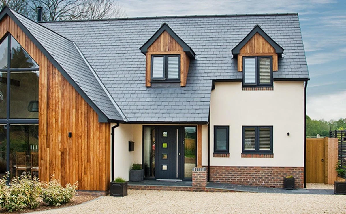 Modern house with a combination of wood, brick, and white exterior walls, with three dormer windows on the roof, large glass window on the left, and a black front door, surrounded by a gravel pathway and landscaped garden.