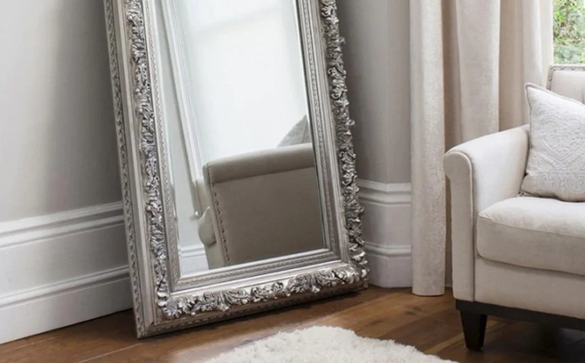 A large ornate silver-framed mirror leaning against a wall, with part of a beige armchair and a window with curtains visible in the room.