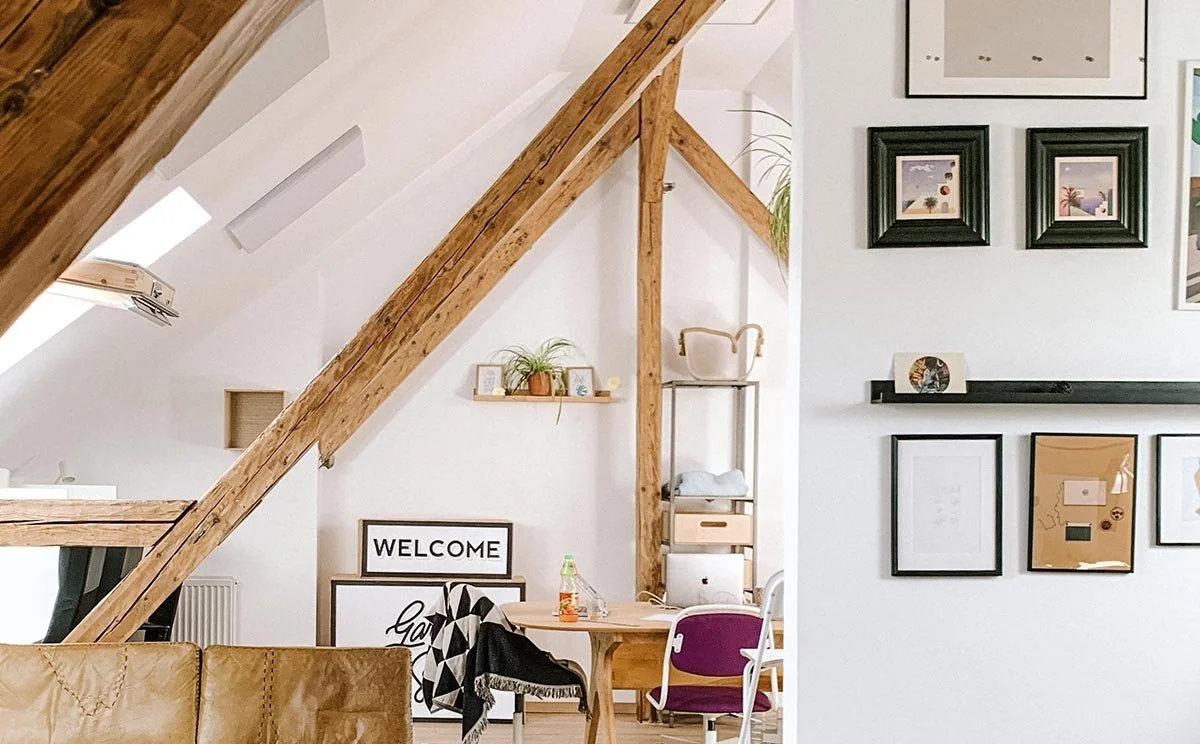 Interior view of a modern living space with wooden beams, a white wall decorated with framed pictures, and a workspace with a desk and chair.