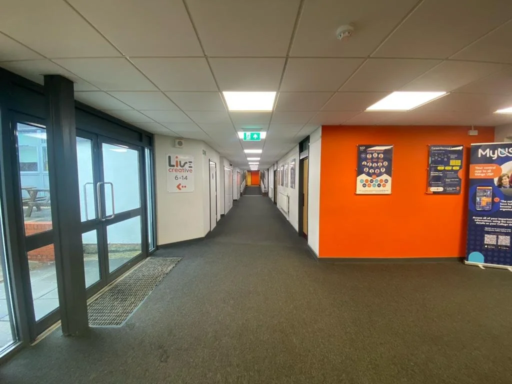 Long indoor corridor with a glass entrance door on the left, white walls with posters, and a bright orange wall on the right side. The ceiling has multiple rectangular fluorescent lights, and there is a dark carpeted floor.
