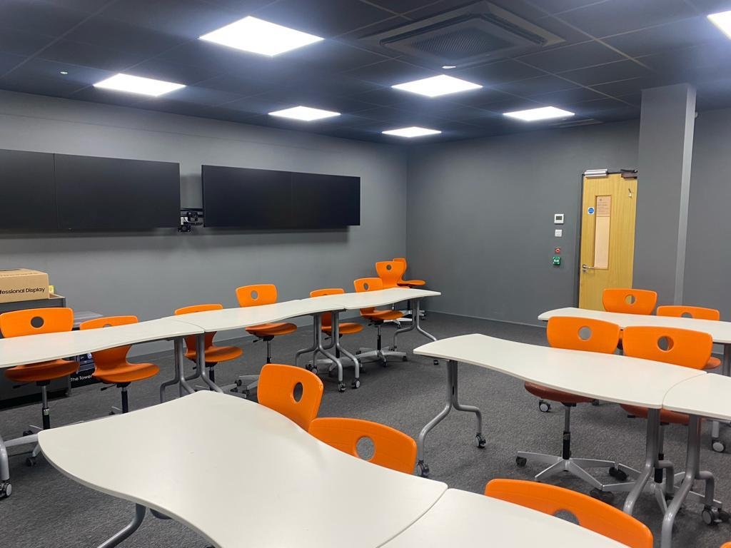 Empty classroom or conference room with orange chairs, white tables, and large display screens on gray walls under bright ceiling lights.